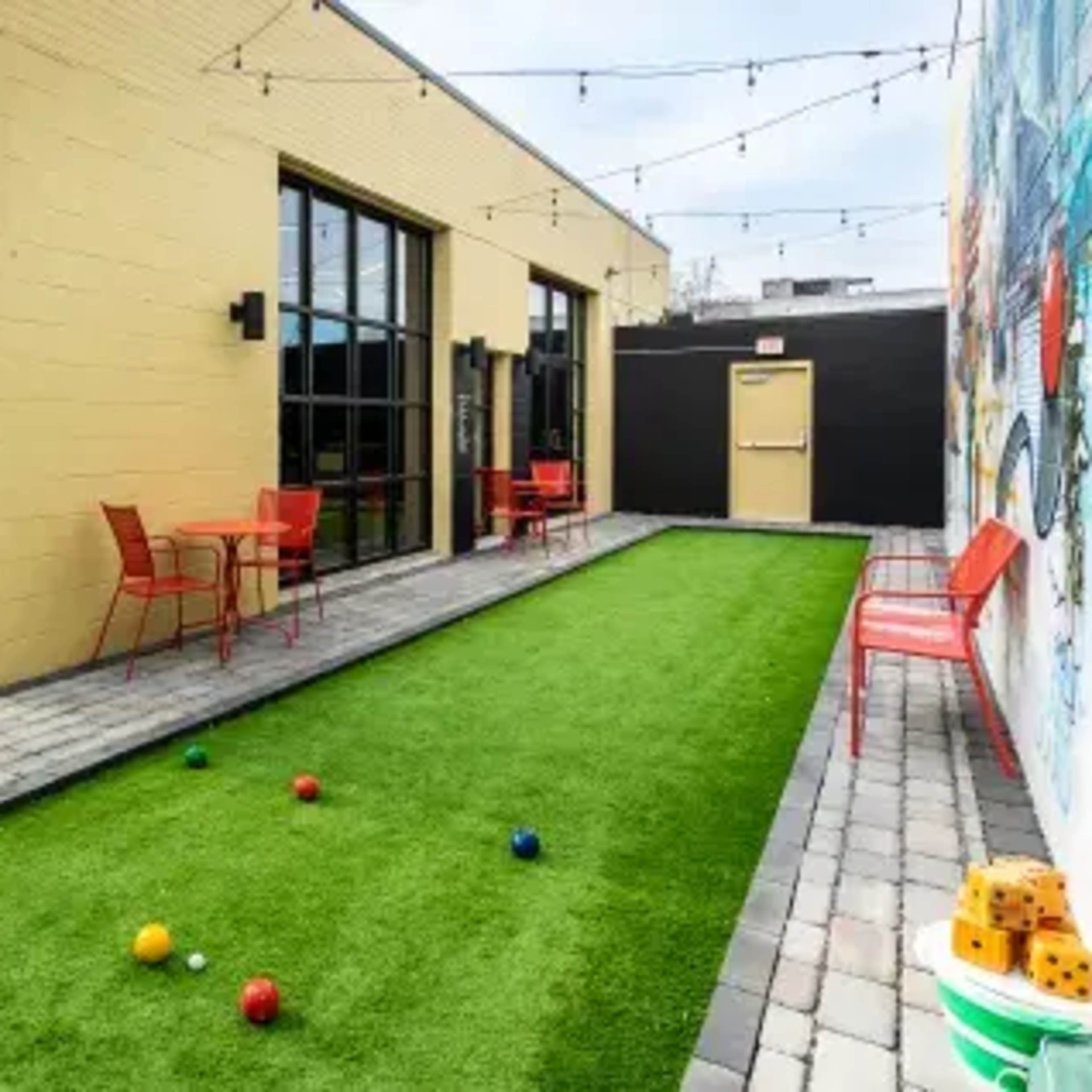 A small outdoor area featuring a grassy surface with colorful balls, adjacent to a wall mural and equipped with red patio furniture.