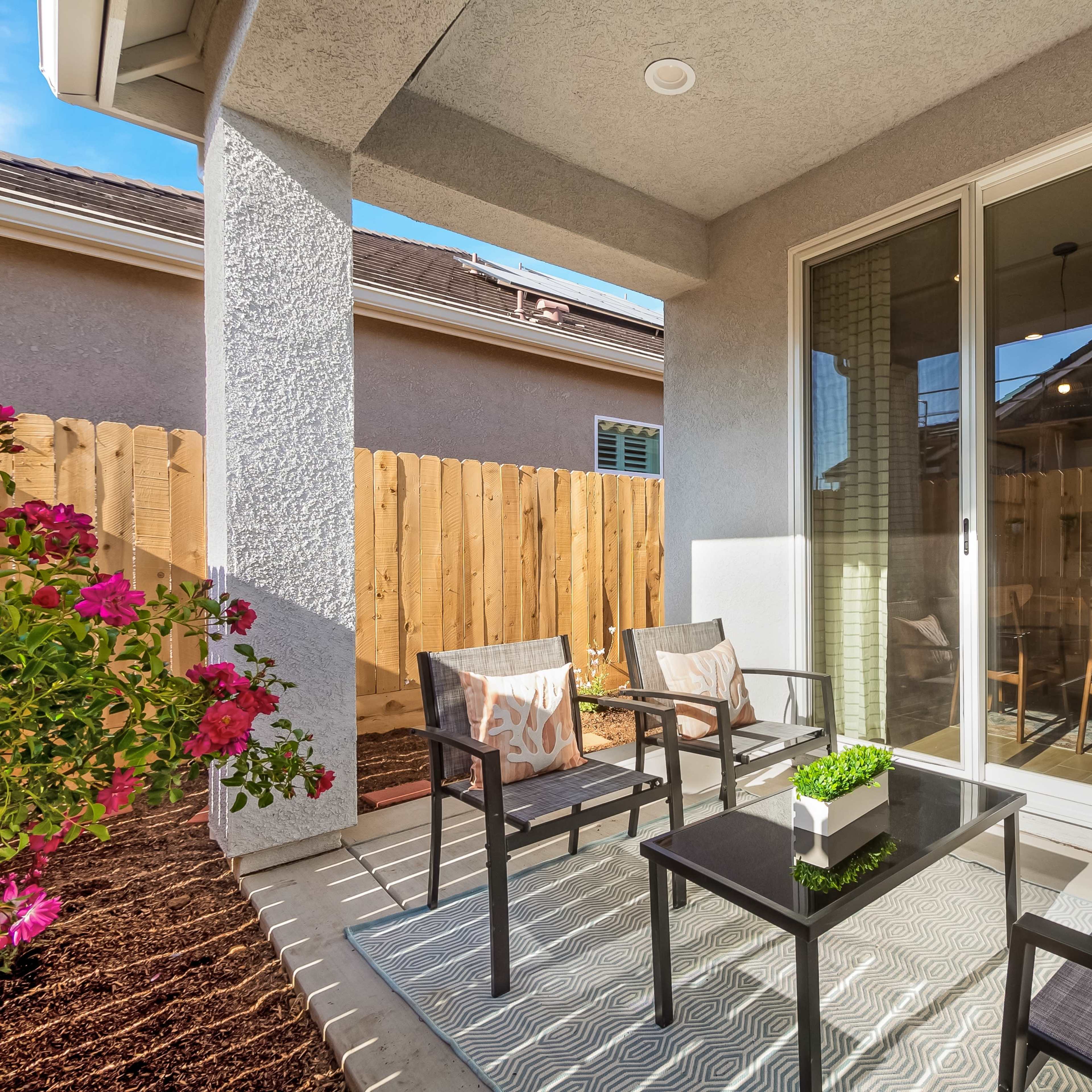 A patio area with two chairs and a table, bordered by a wooden fence and featuring flowering plants.