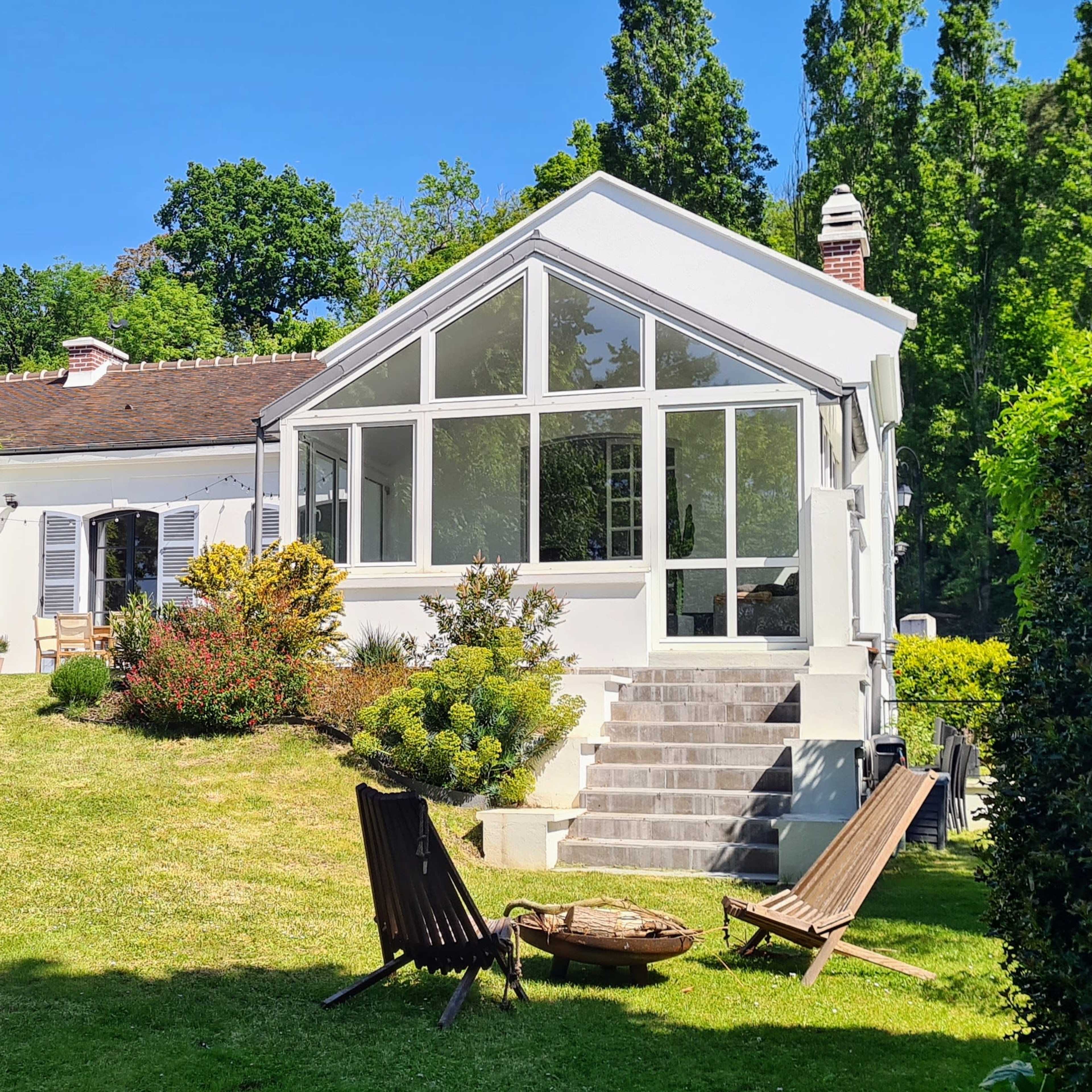 A modern house with a large glass-enclosed patio sits on a green hillside surrounded by trees and landscaped gardens.