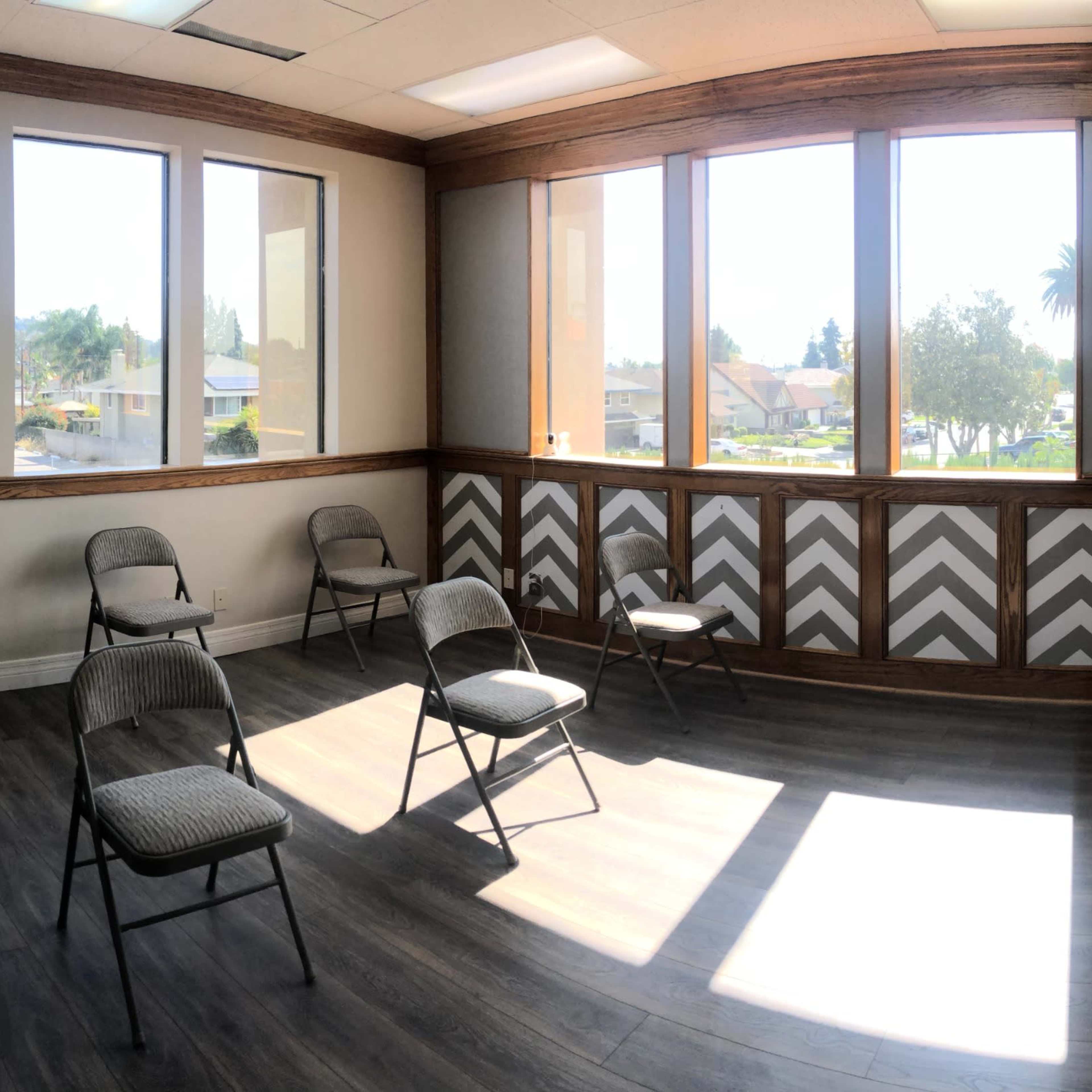 A spacious room with several folding chairs arranged on a wood-style floor, large windows providing natural light, and a television mounted on the wall.
