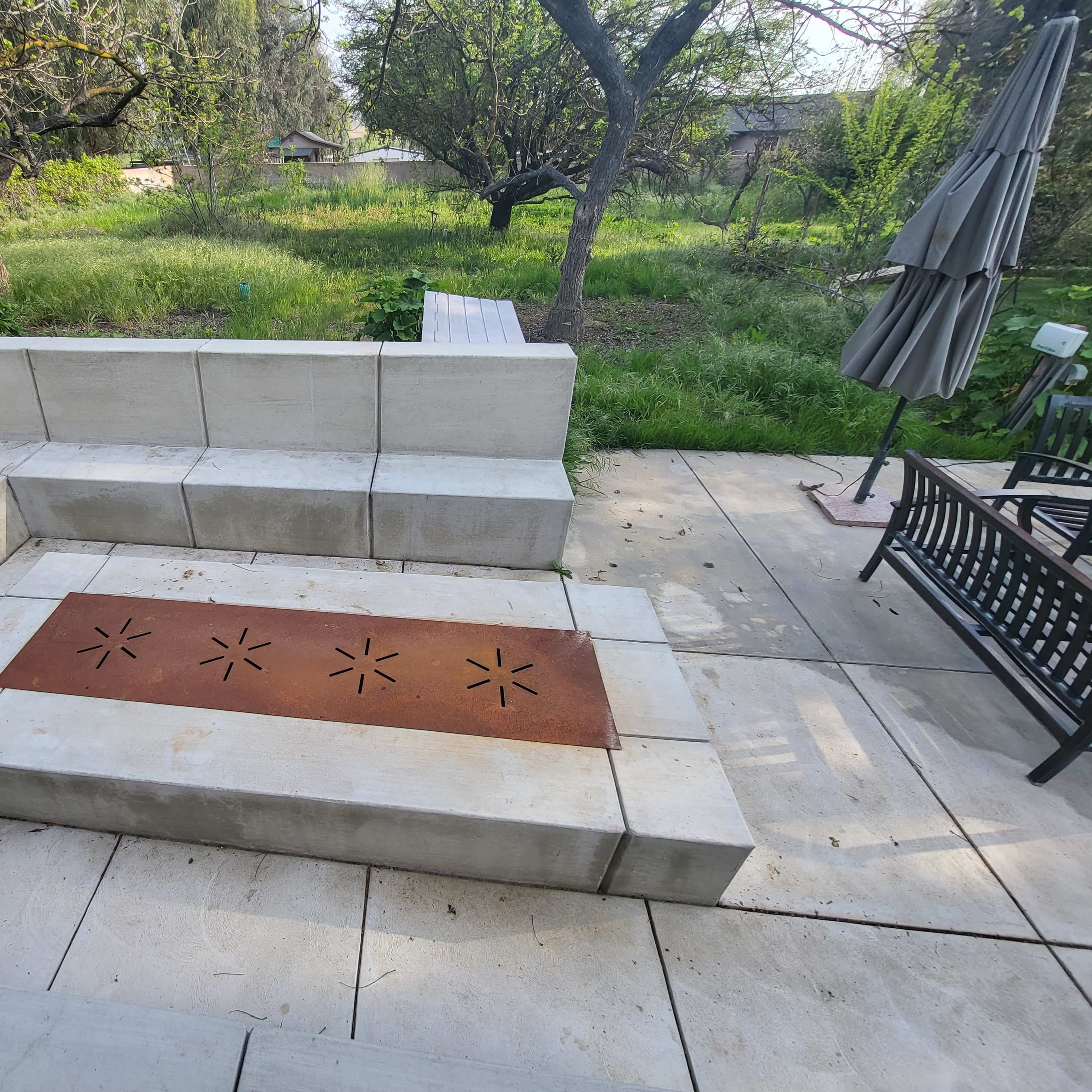 A stone patio area with a metal fire pit and surrounding seating, set in a green backyard with trees and grass.