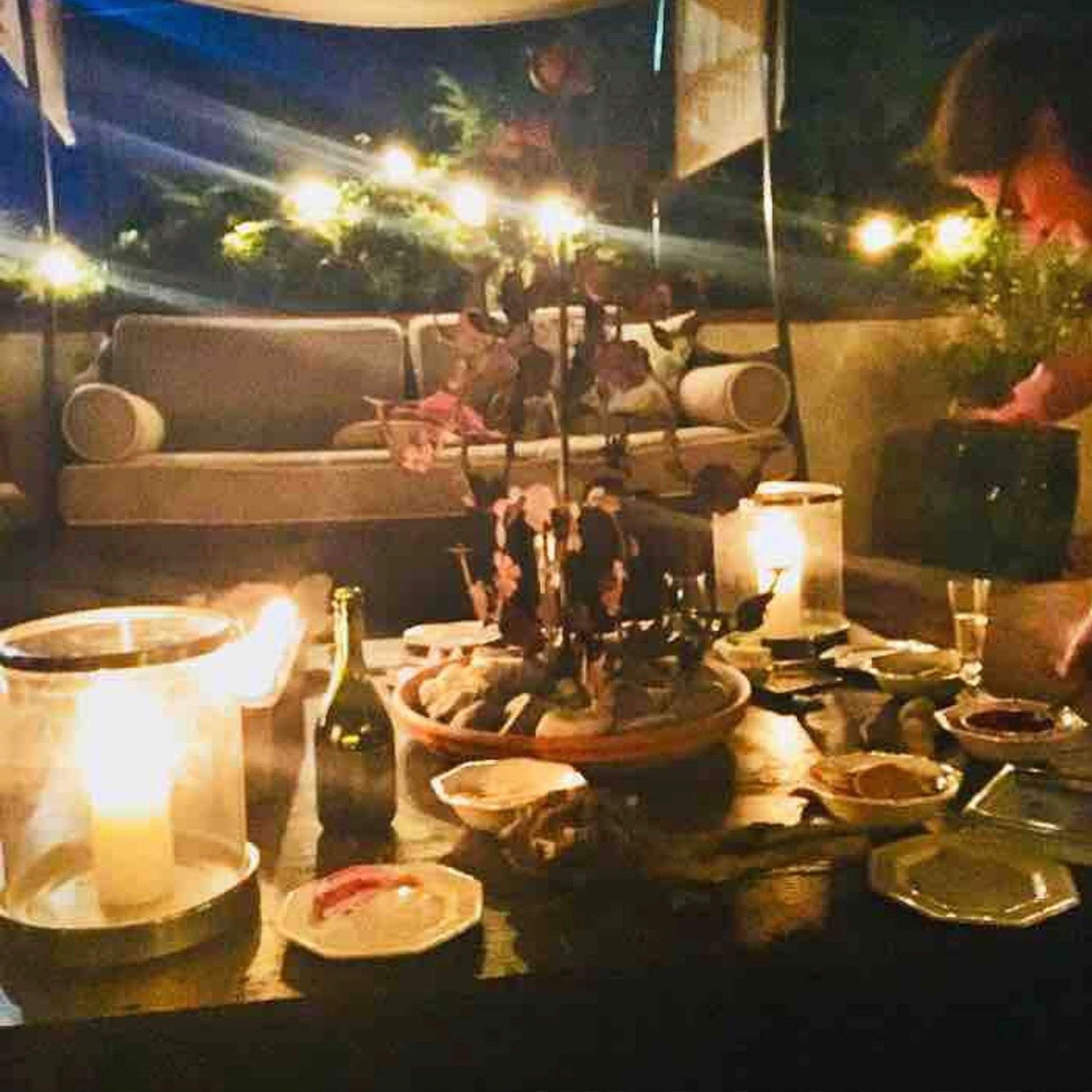 A cozy outdoor setting features a table with plates of food, candles, and a person preparing items nearby, with soft lighting from string lights in the background.