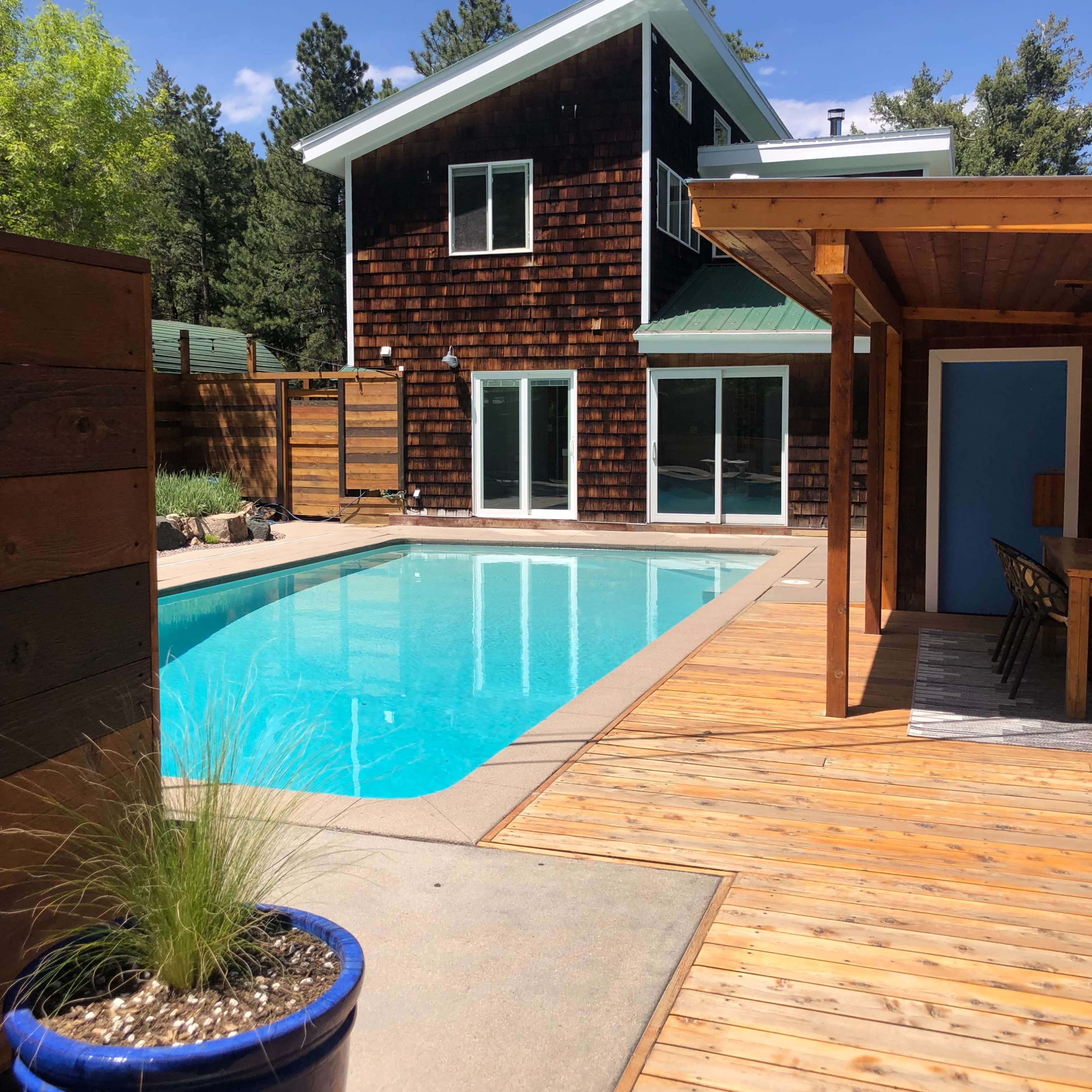A modern house with a wooden exterior, a swimming pool in the foreground, and a deck area that includes a dining space.