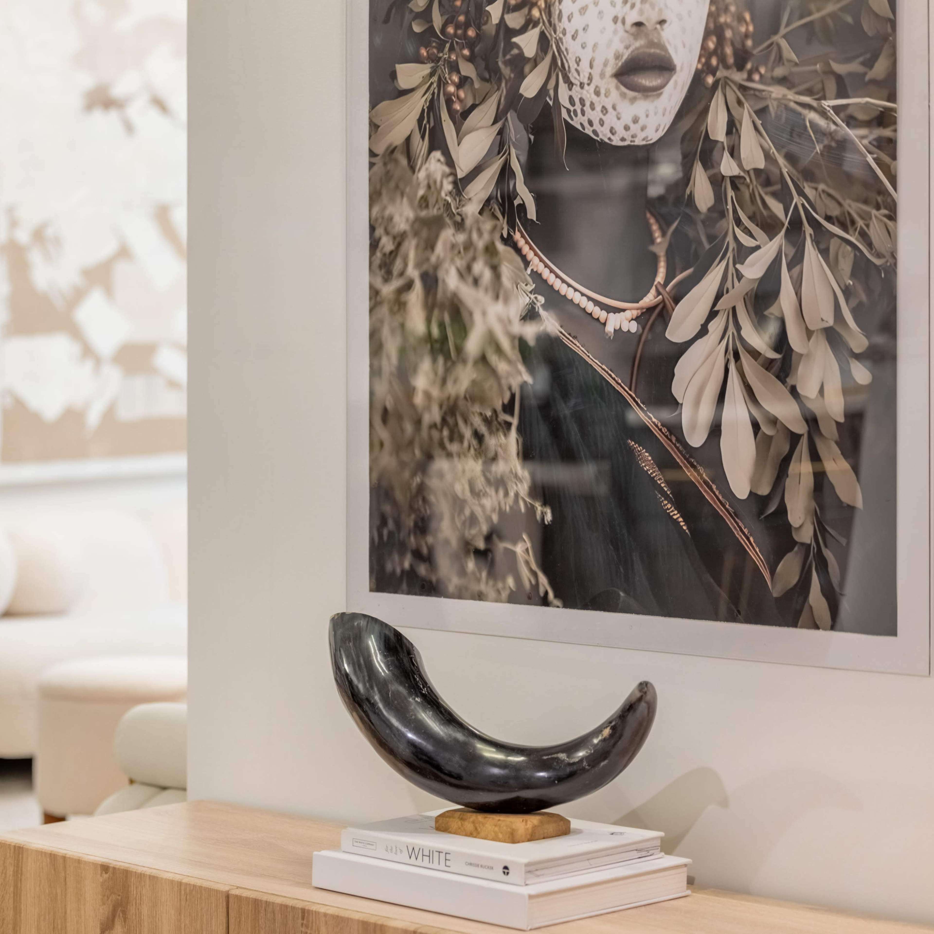 A curved black sculpture sits on a wooden tabletop in the foreground, with a photograph of a person adorned with greenery on the wall behind it.