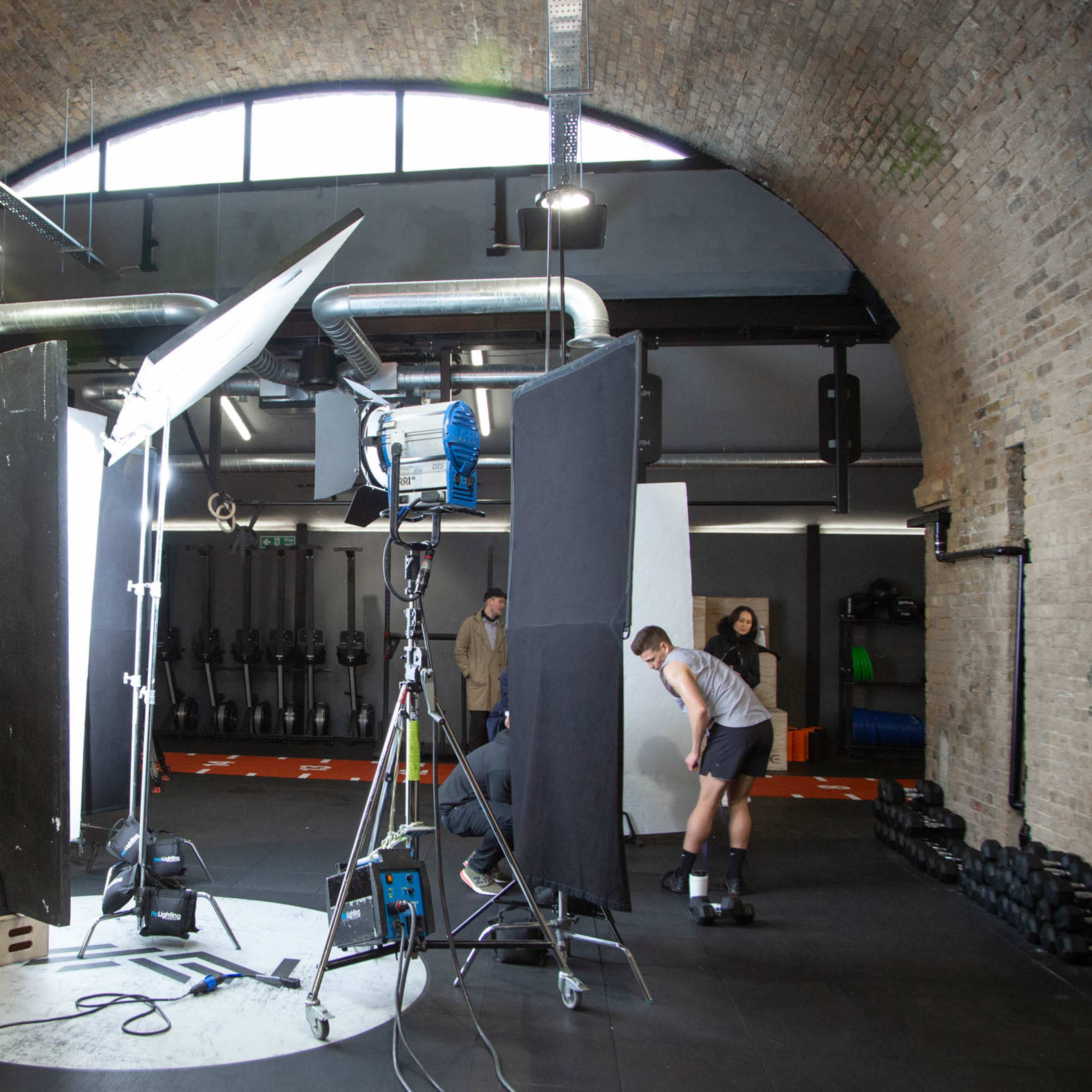 A film studio setup features a large light source, black and white backdrops, and crew members preparing equipment in an arched brick room.