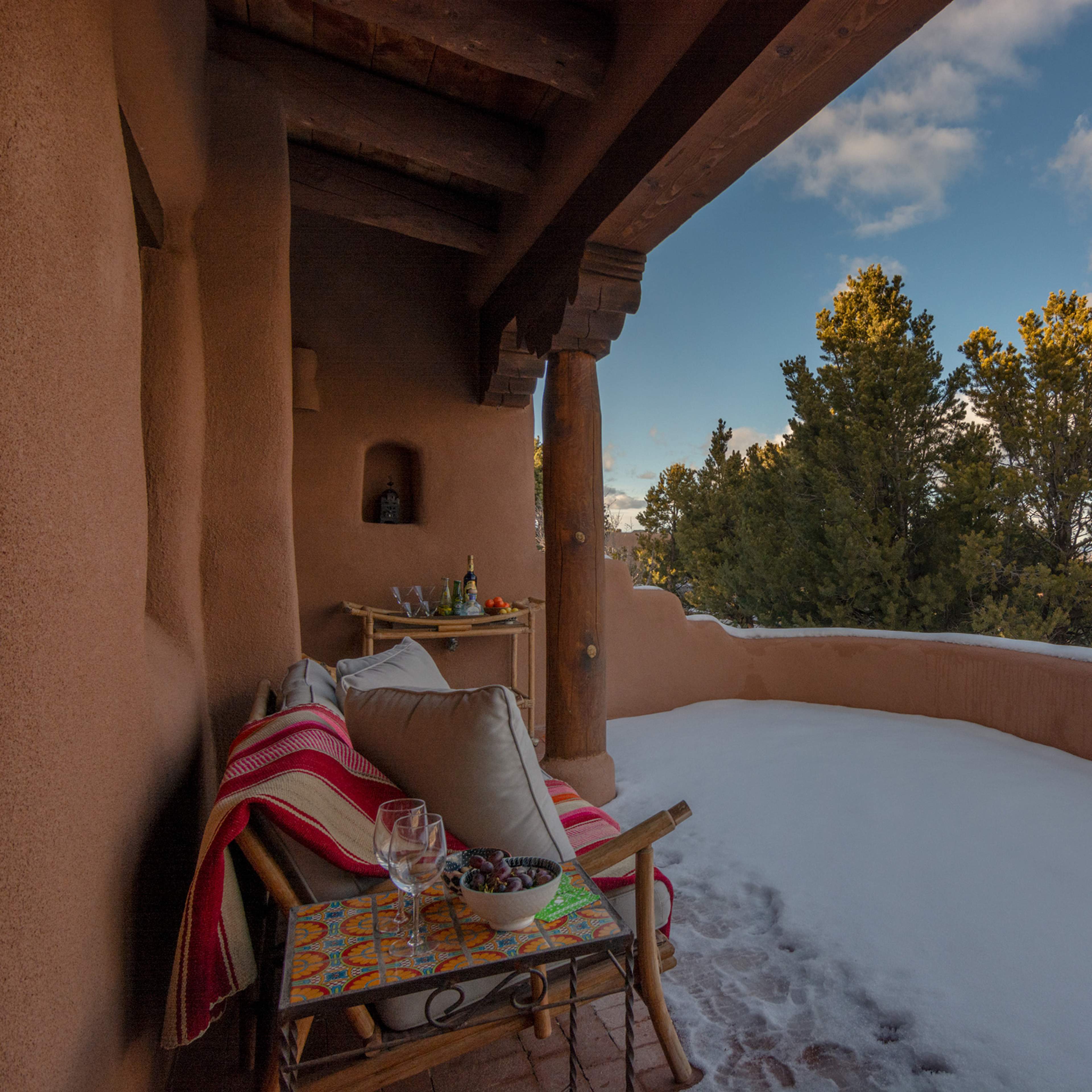 A cozy outdoor seating area features a woven chair and a small table on a snow-covered balcony, surrounded by trees and mountains under a cloudy sky.