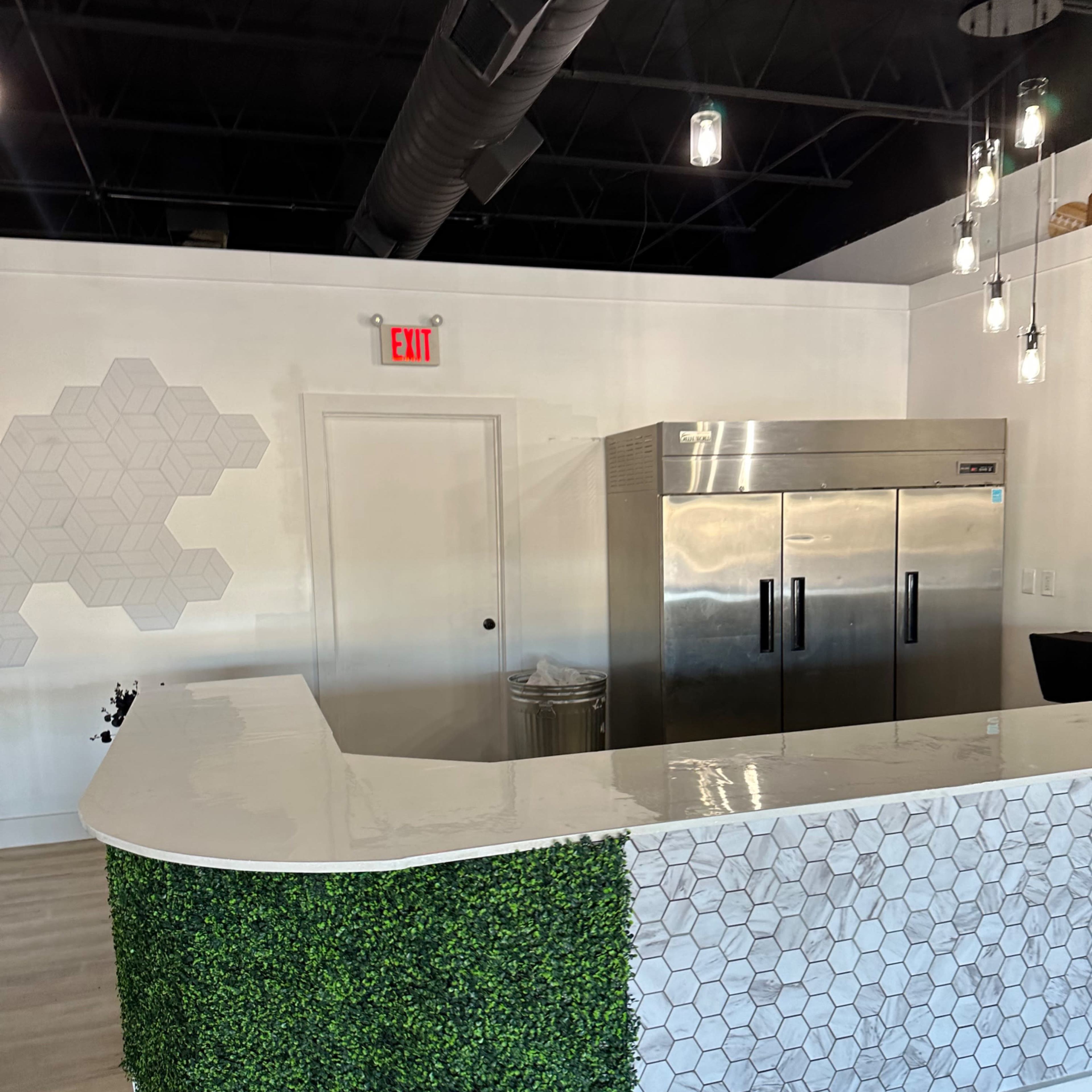 A modern reception area featuring a counter with a geometric design, a stainless steel refrigerator, and green hedging decor on the front.