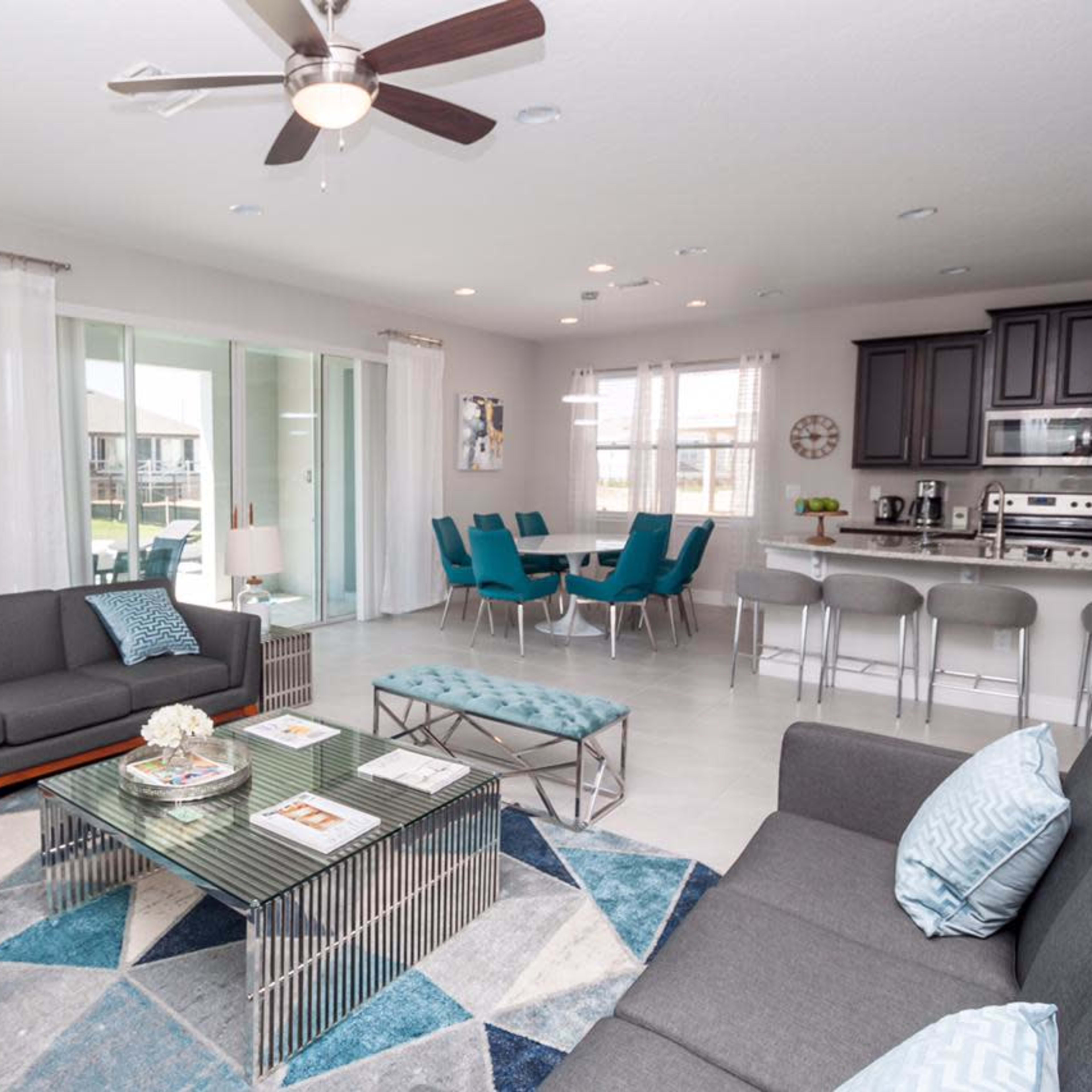 A modern living room with two sofas, a coffee table, a decorative area rug, and a view into a dining area with teal chairs and a kitchen.
