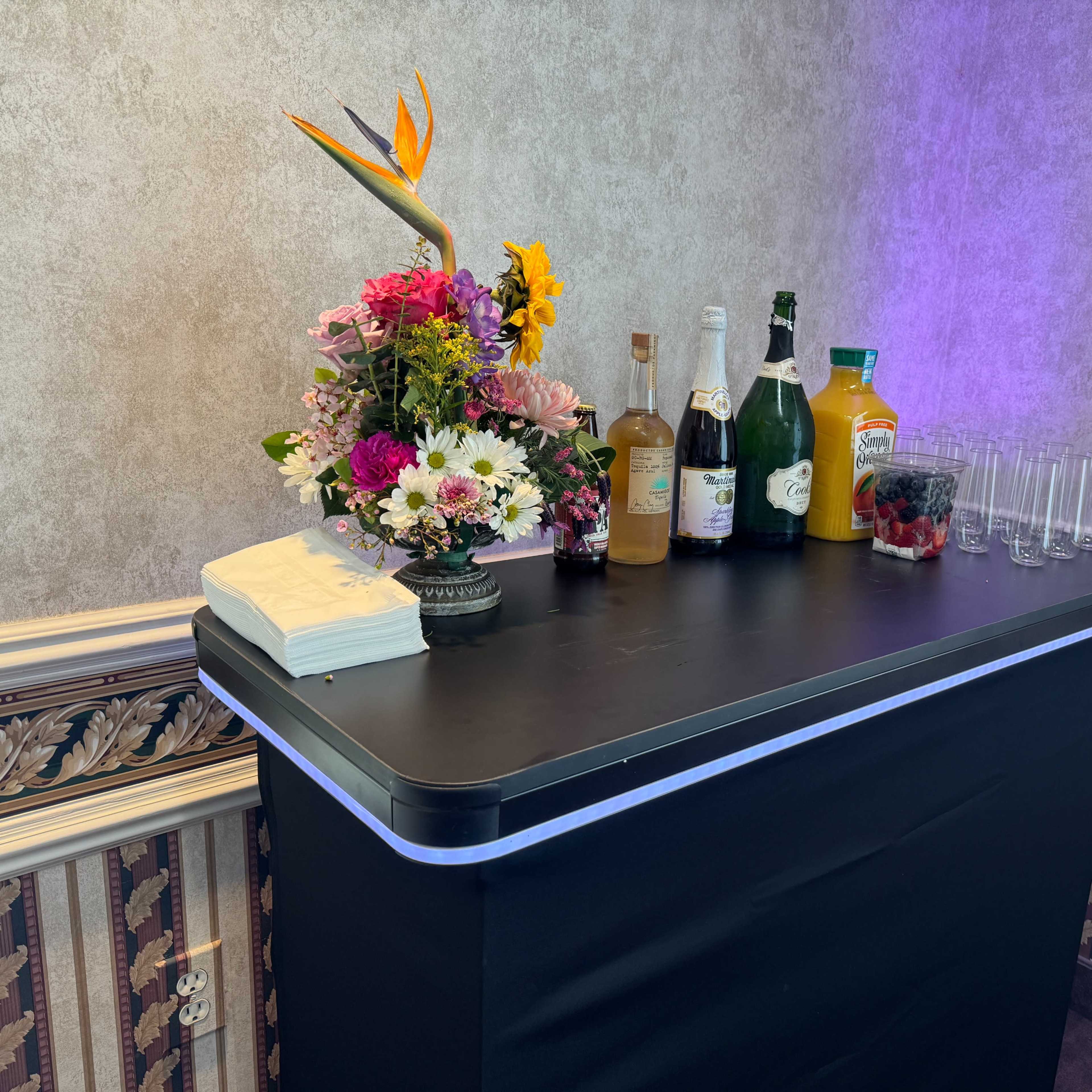 A bar setup is displayed against a textured wall, featuring various bottles of drinks, a floral arrangement, and several empty glasses.