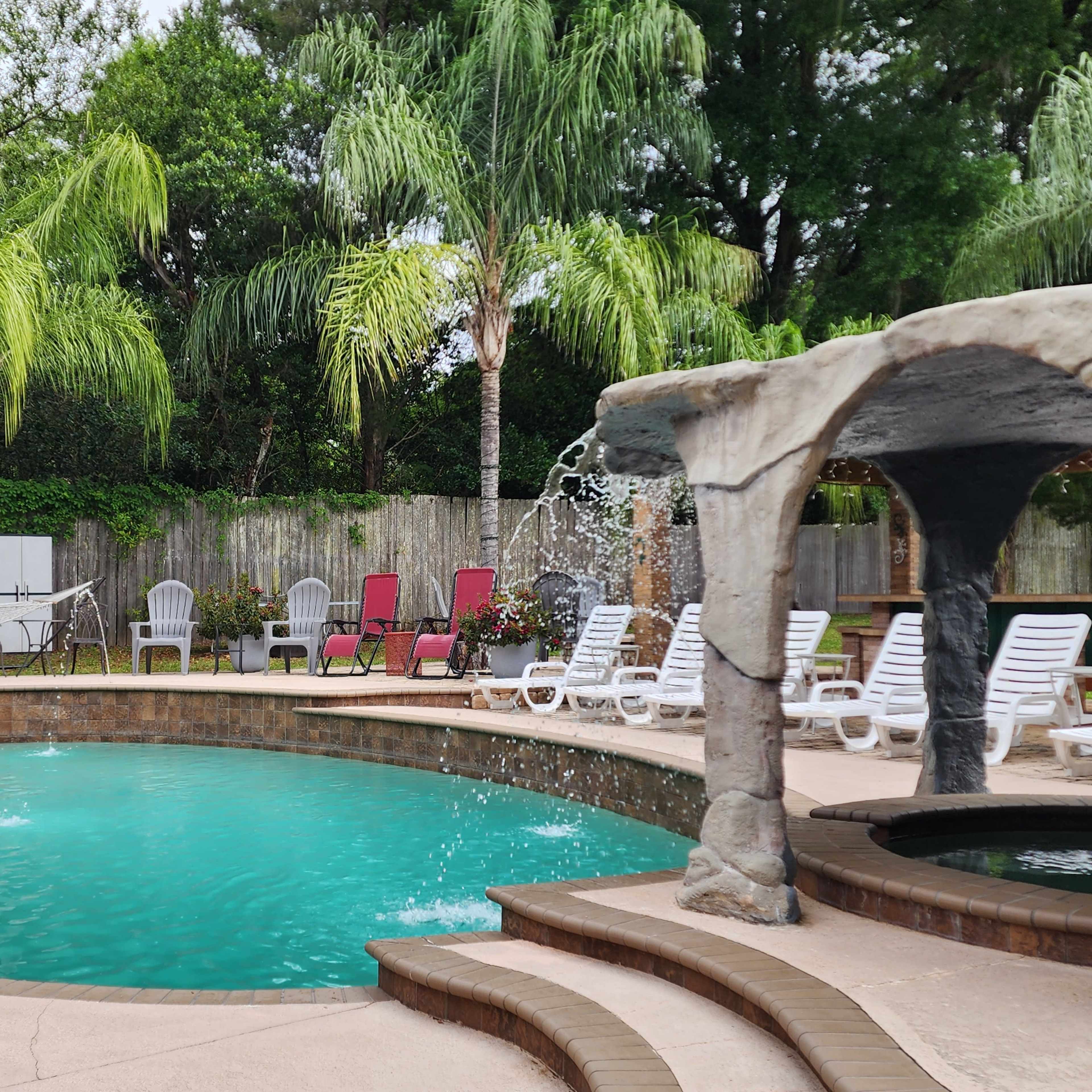 A swimming pool surrounded by lounge chairs and palm trees, with a rock fountain and a beach ball floating in the water.