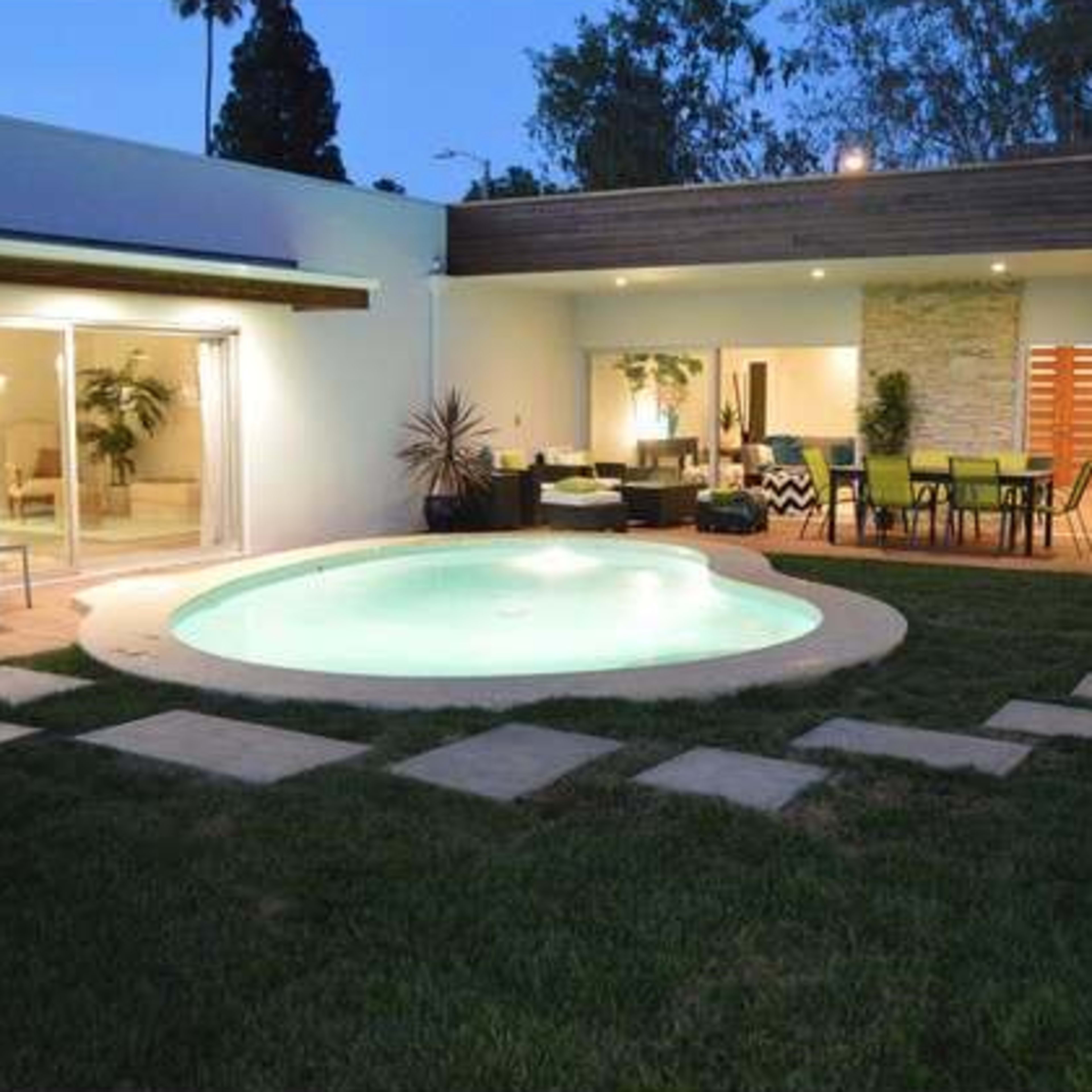 A modern home with a circular pool surrounded by grass and stone pathways, featuring sliding glass doors that open to an outdoor seating area.