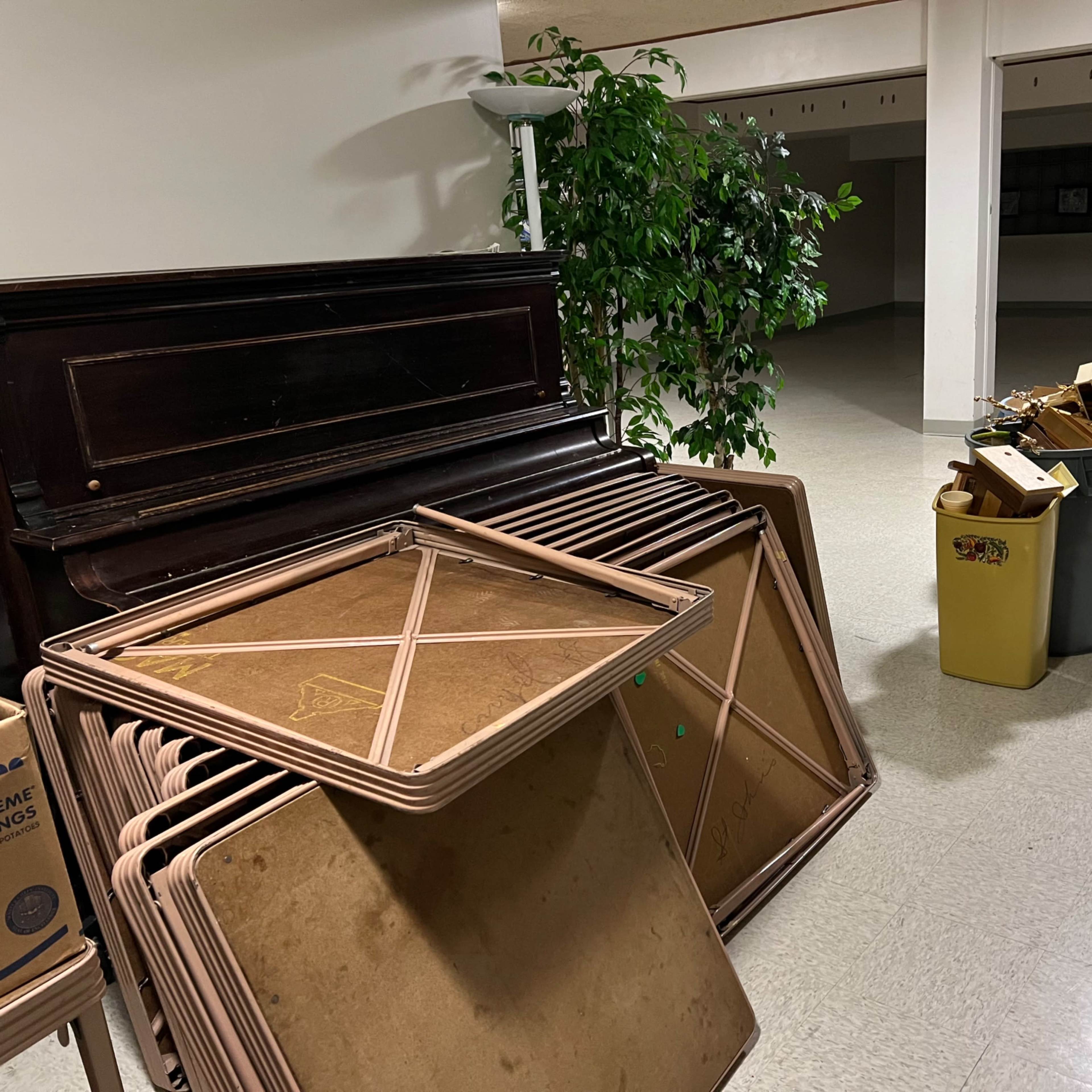 A room with stacked folding tables, a piano, a potted plant, and a collection of boxes and trash cans on the floor.