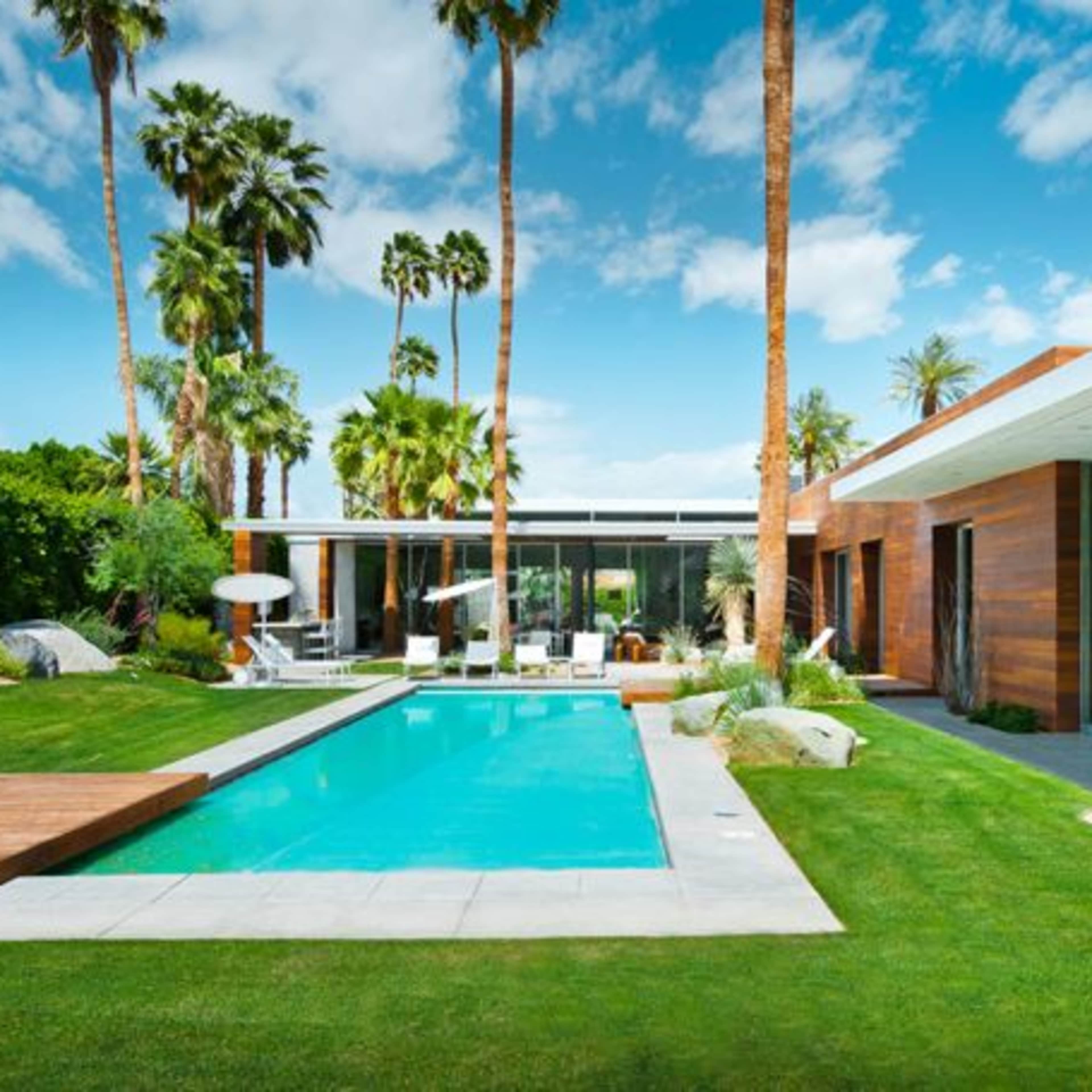 A modern home with large windows and a swimming pool is surrounded by palm trees and well-manicured grass.