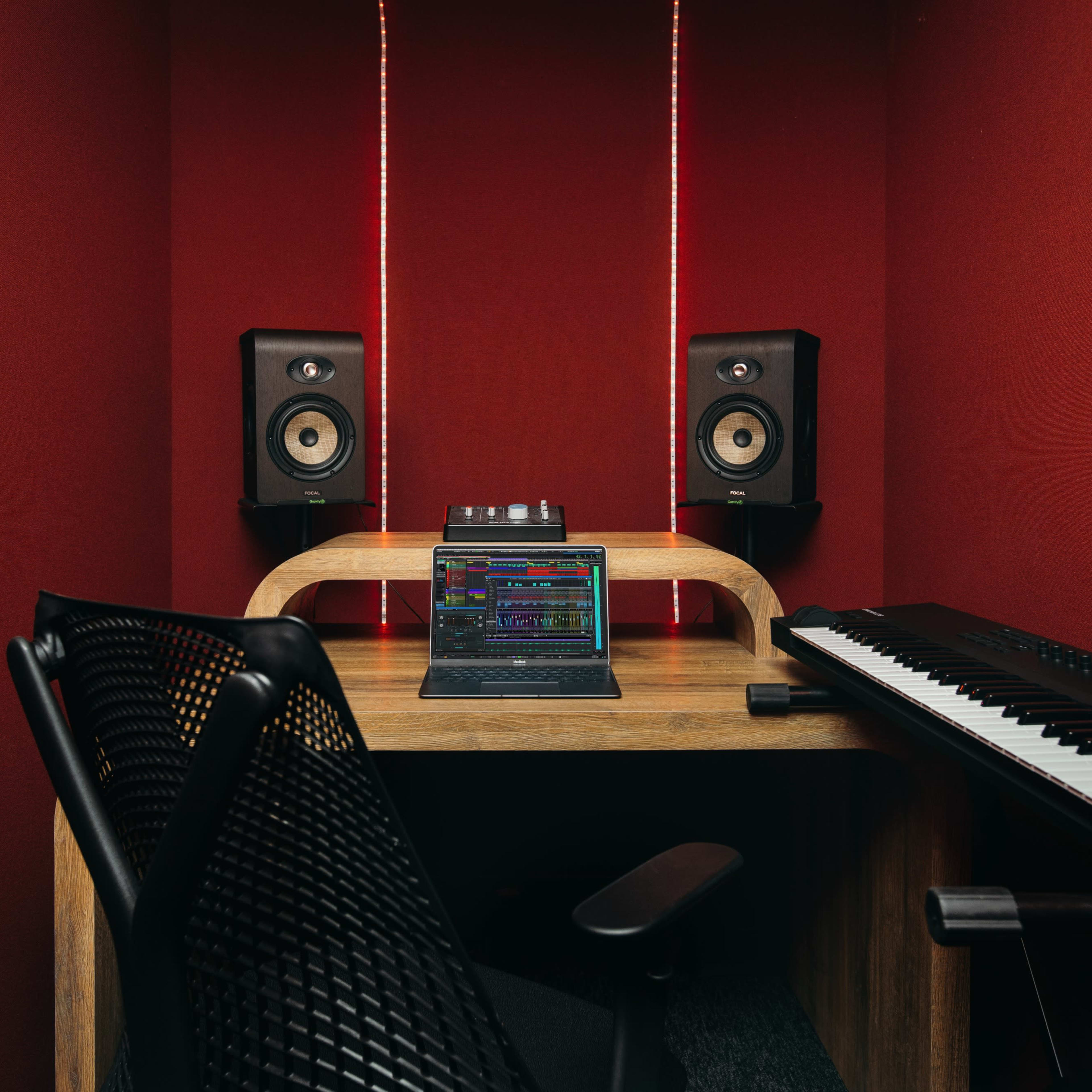 A soundproof studio corner features a wooden desk with a laptop displaying audio software, flanked by studio monitors and a keyboard, set against a red wall.