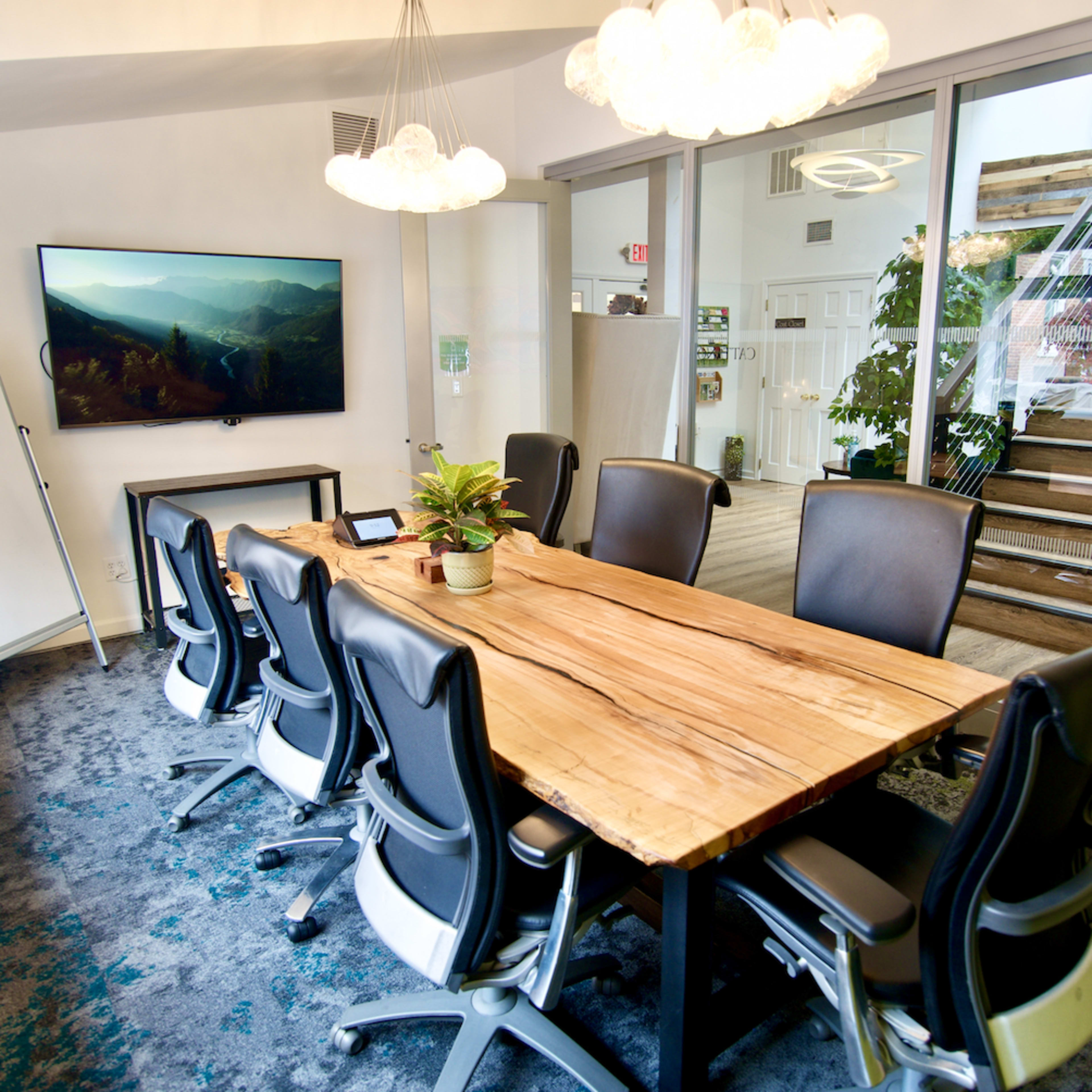 A modern conference room with a large wooden table, ergonomic chairs, a television mounted on the wall, and a whiteboard, alongside glass doors leading to a staircase and greenery.