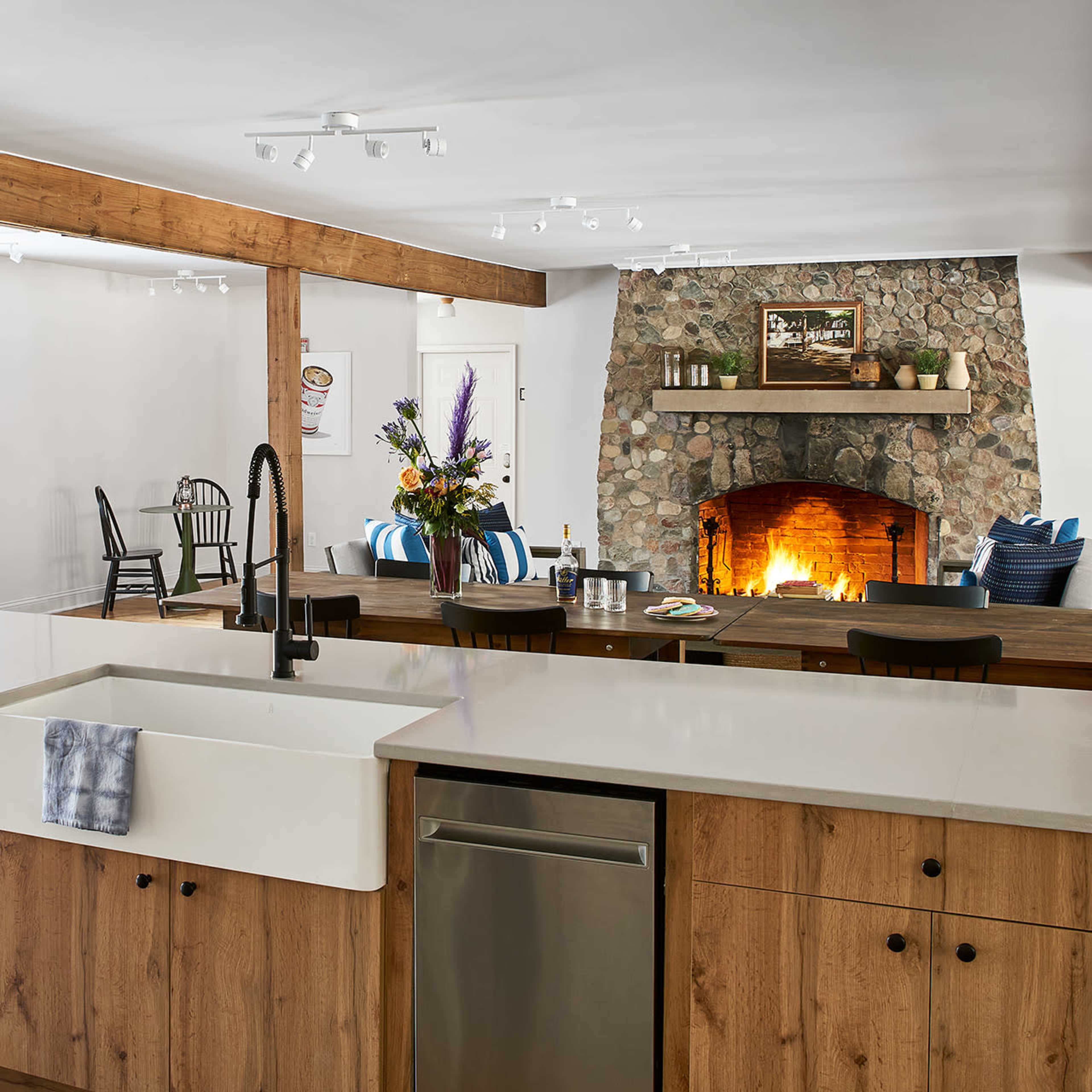A modern kitchen features a large farmhouse sink and wooden cabinets, opening into a spacious dining area with a stone fireplace.