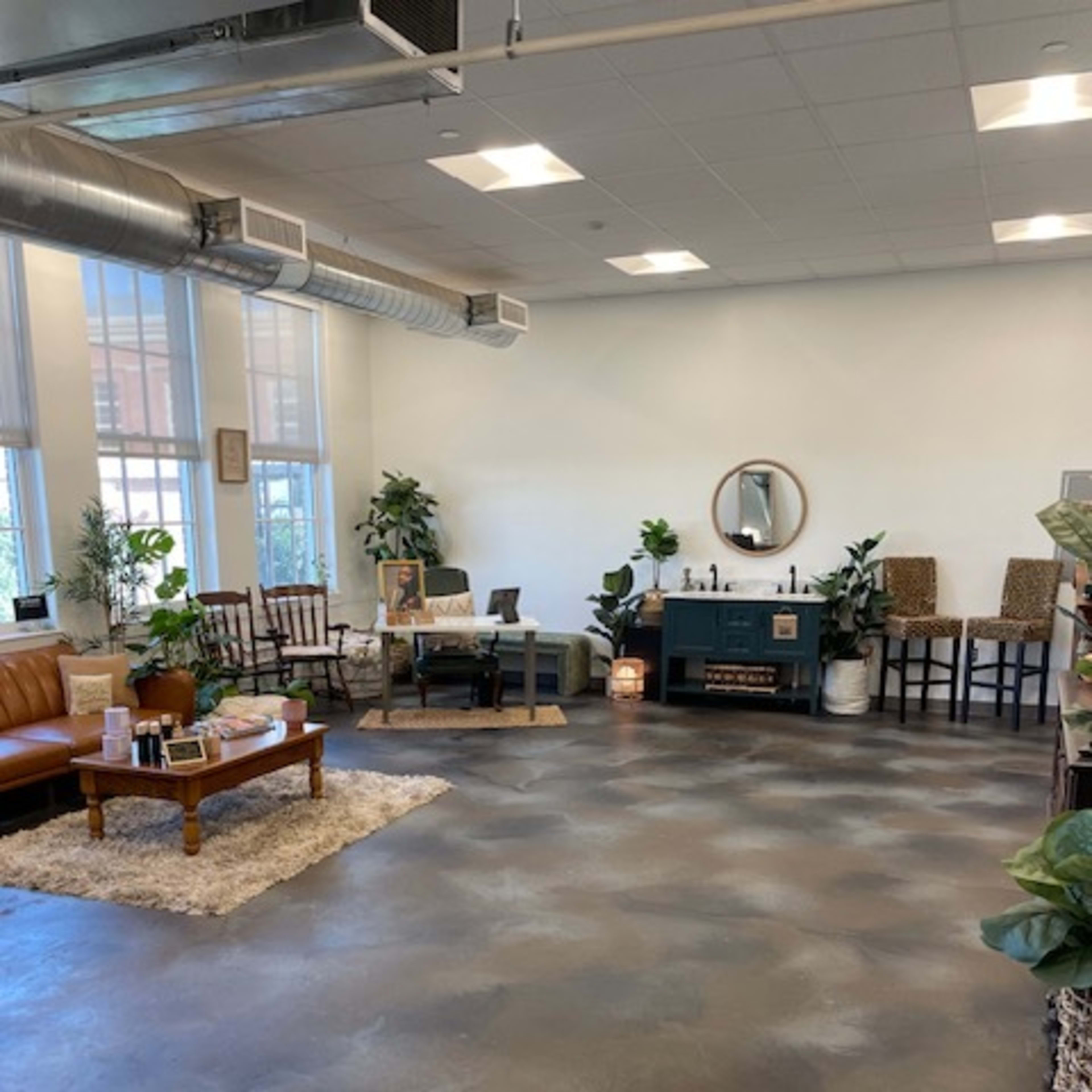 A spacious room with a concrete floor, furnished with a brown leather couch, wooden chairs, a coffee table, and a dark cabinet, surrounded by indoor plants.