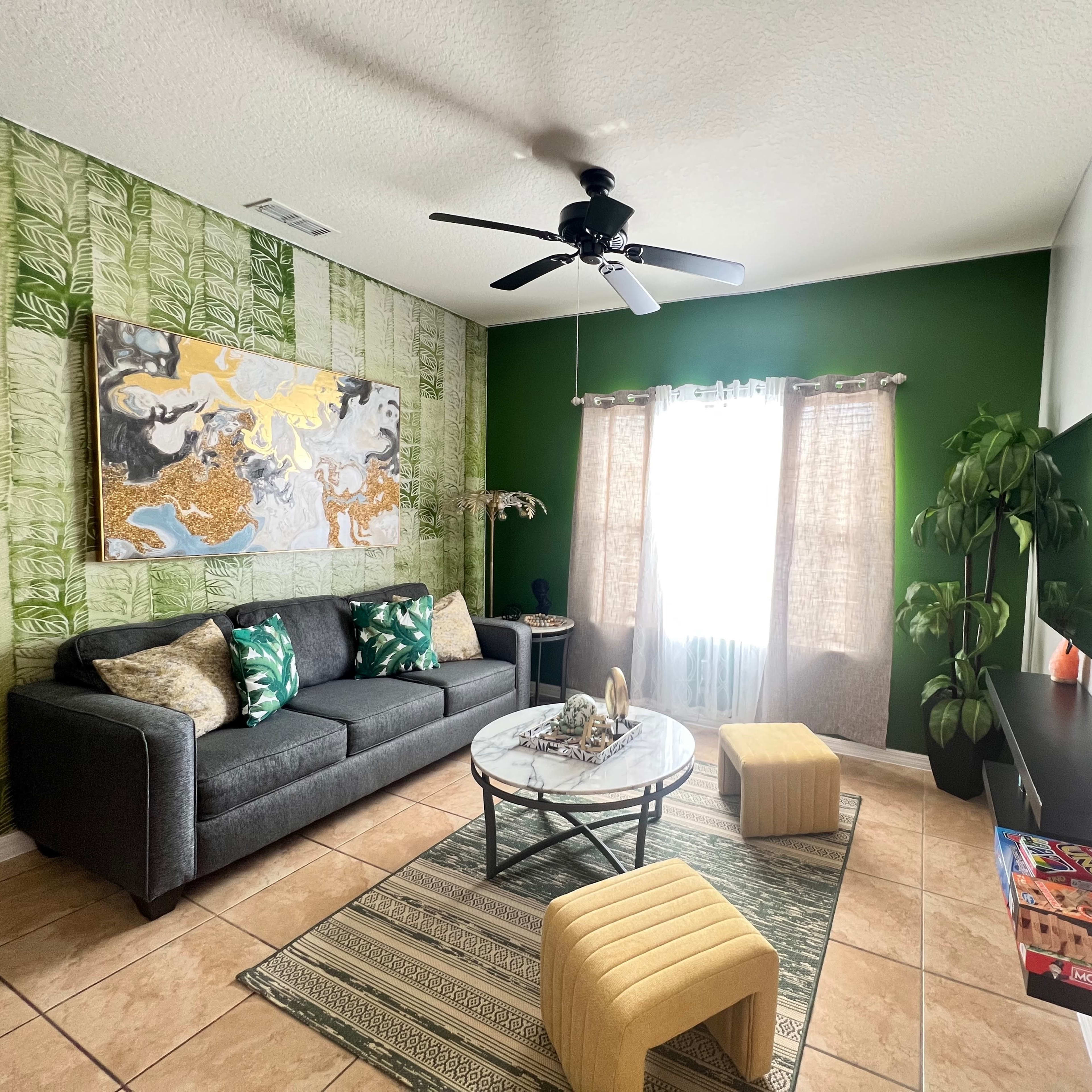 The living room features a gray sofa, a round coffee table, a large colorful wall painting, and a green accent wall with natural light coming through the sheer curtains.