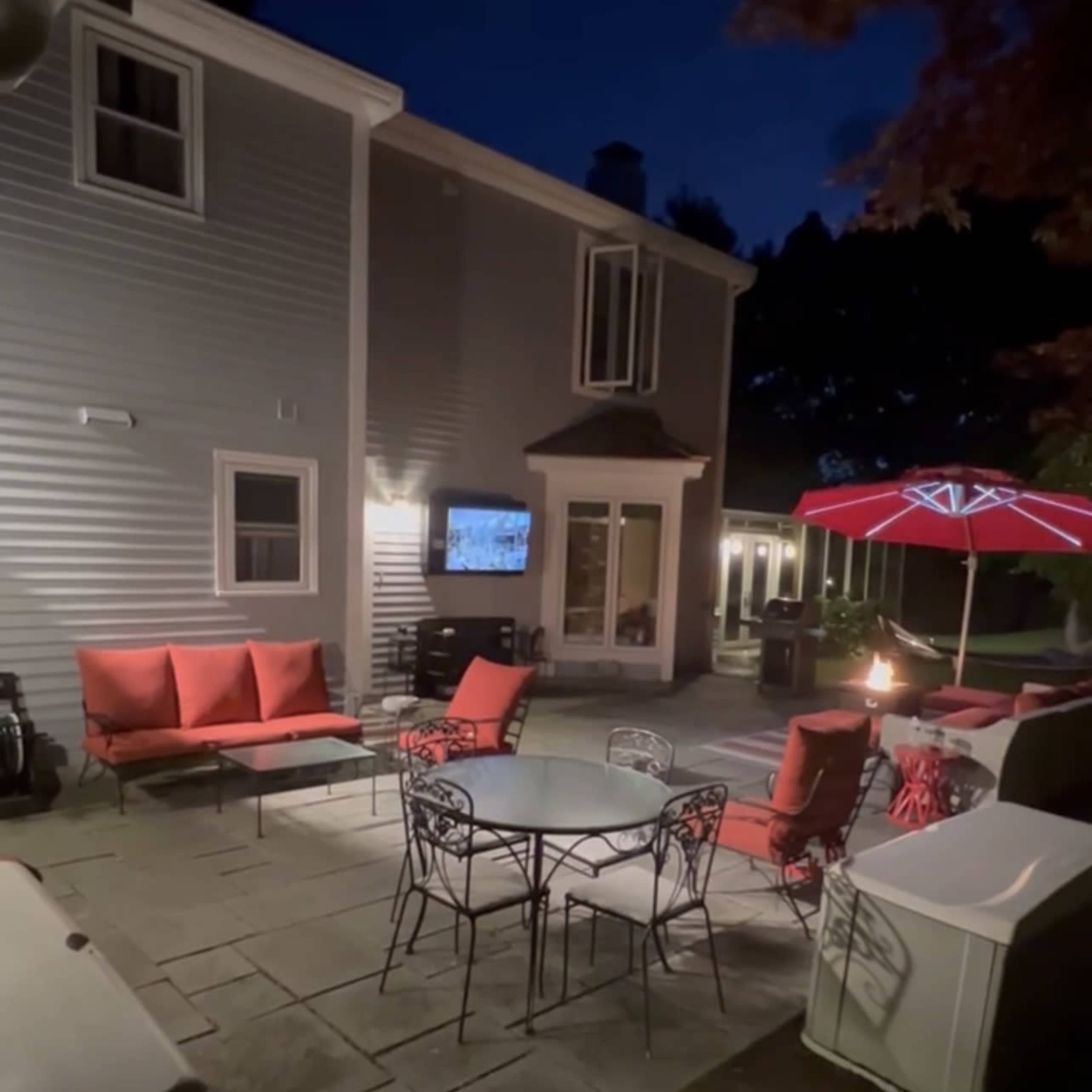 A backyard patio at night with a table and chairs, a fire pit, and a large umbrella over a seating area, alongside a house.
