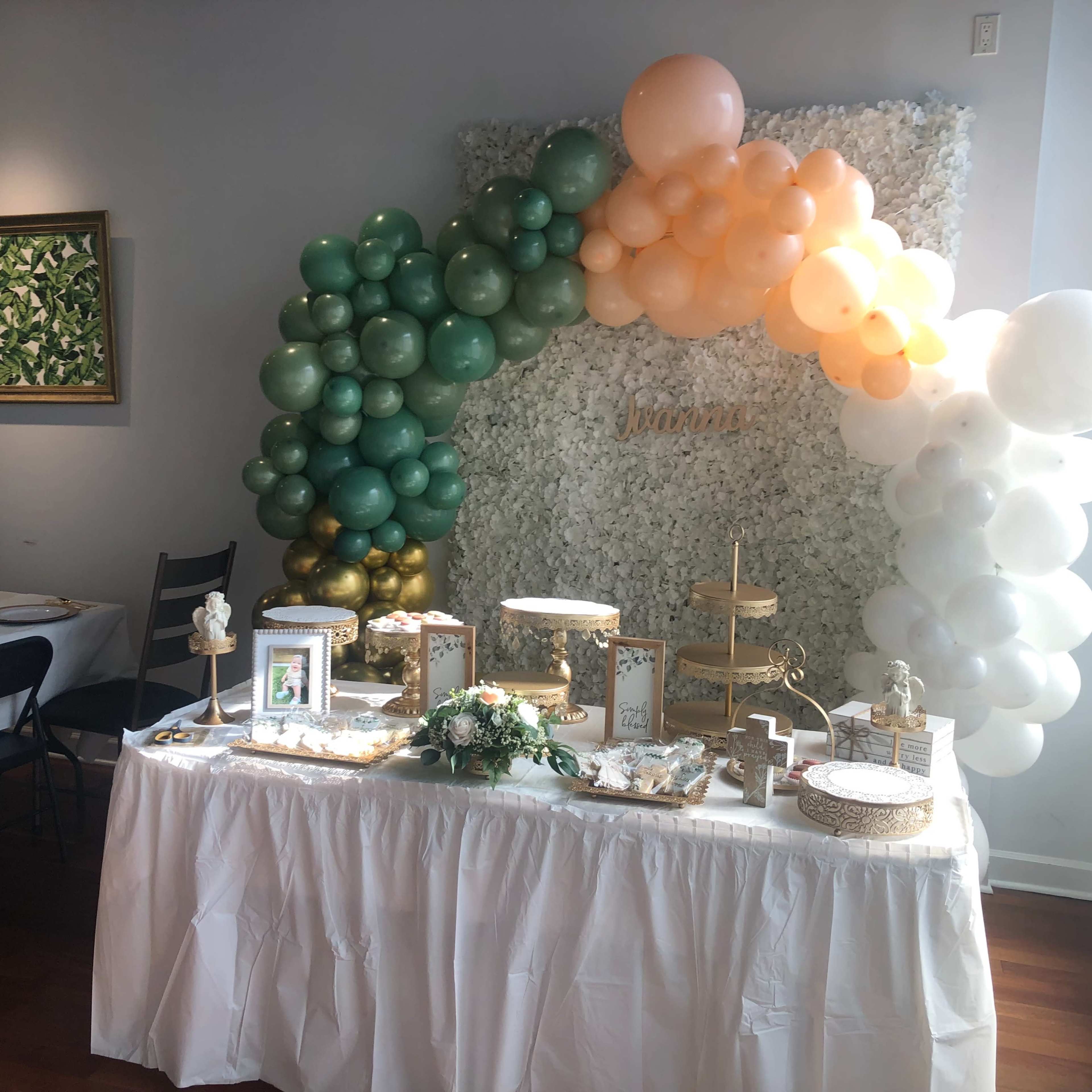 A festive dessert table is decorated with a variety of treats and adorned with a colorful balloon arch against a floral backdrop.