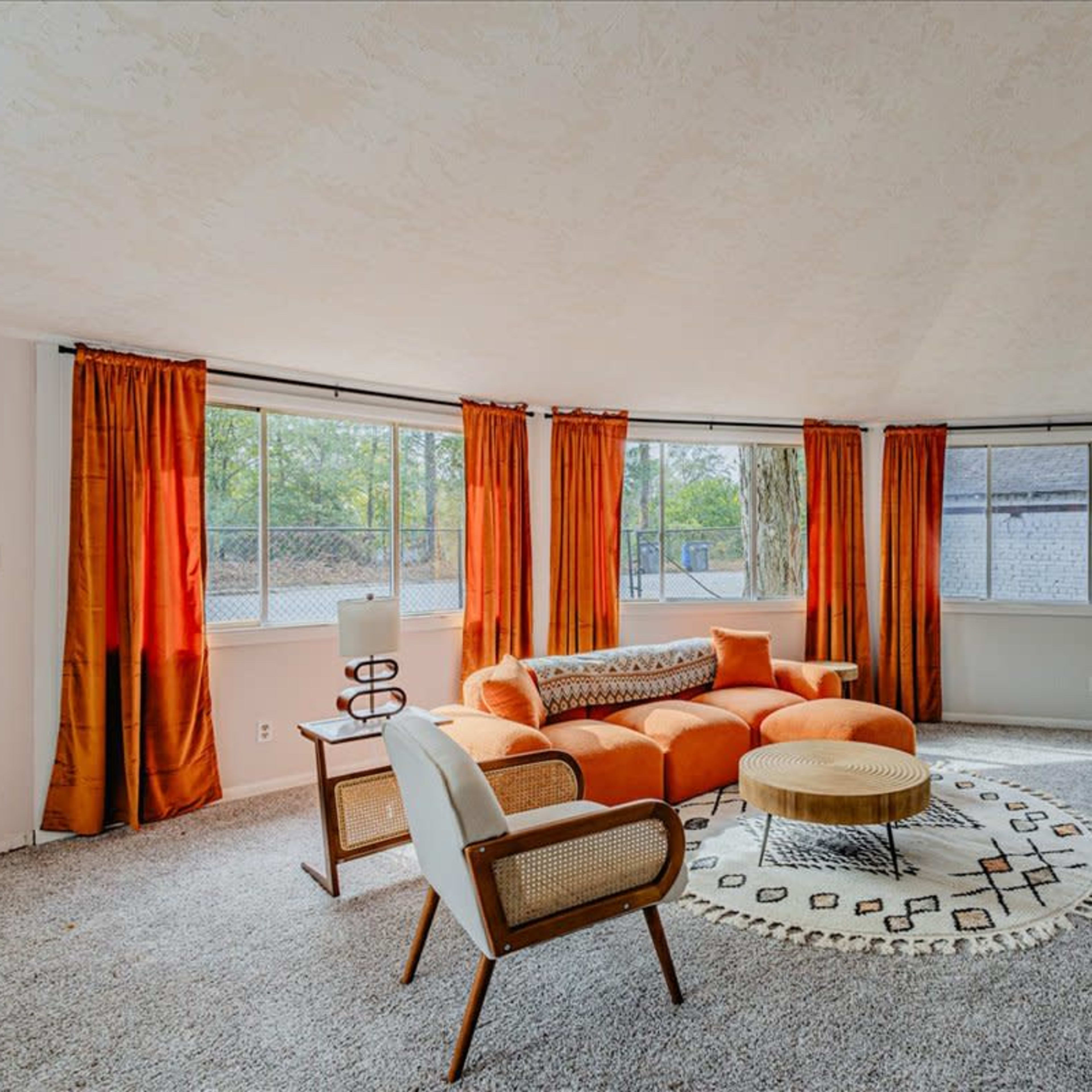 The room features a rounded design with orange curtains, a circular area rug, a modern orange sofa, and two chairs arranged around a coffee table.
