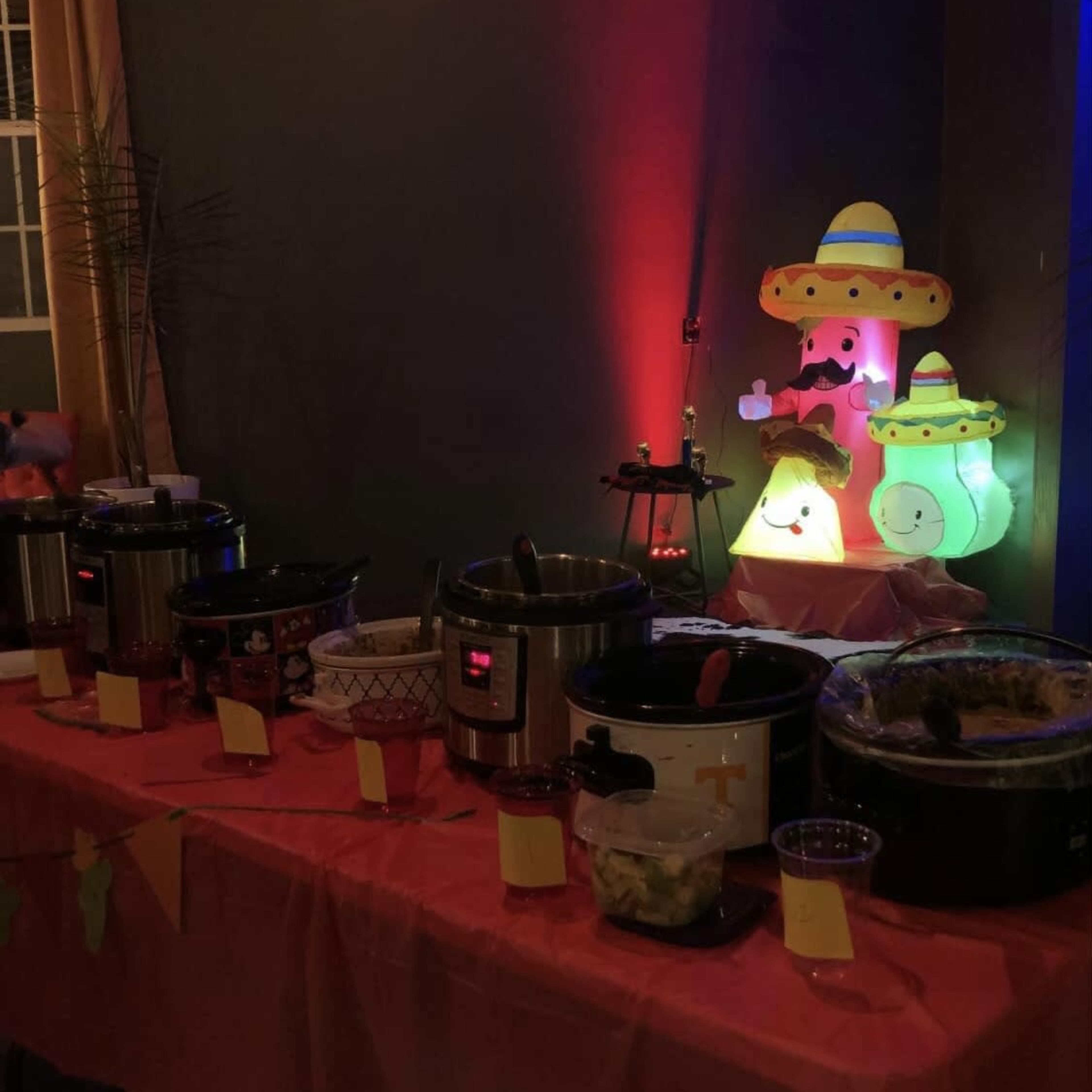 A table decorated with a colorful tablecloth displays several slow cookers and a festive inflatable figure wearing a sombrero.