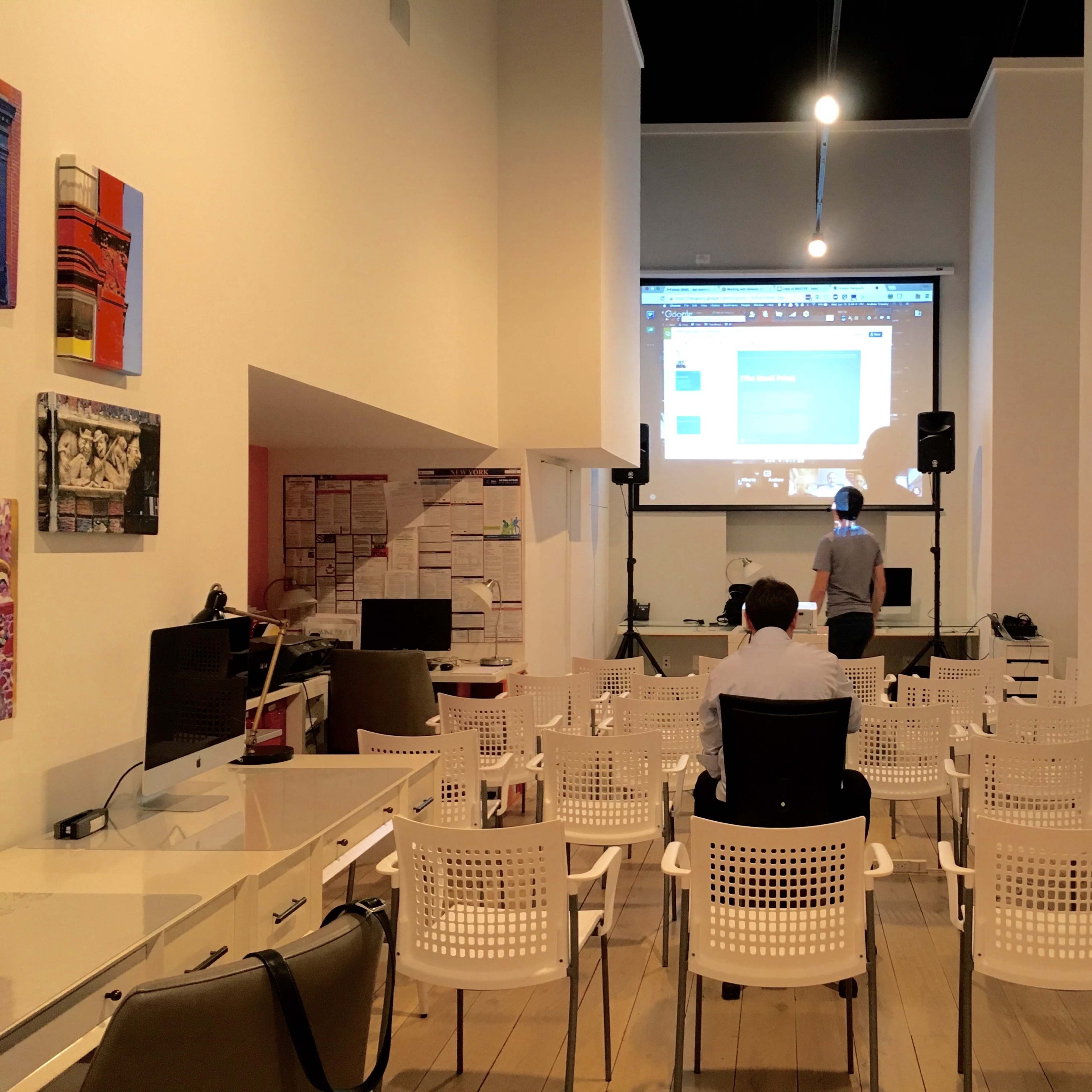 A person stands in front of a projector screen in a room filled with empty white chairs and modern workstations.