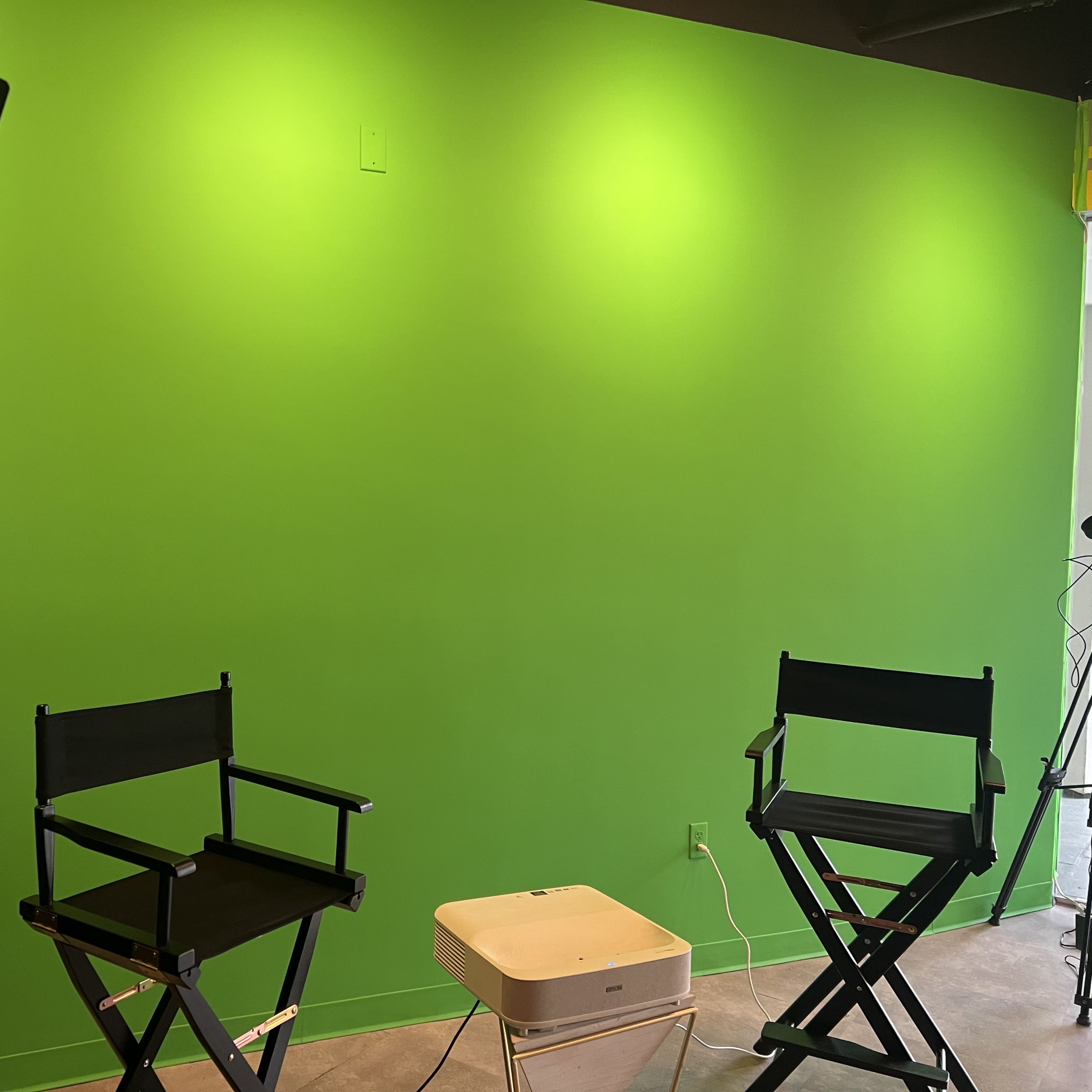 A green screen setup with two director's chairs and a projector in front of a brightly lit green wall.
