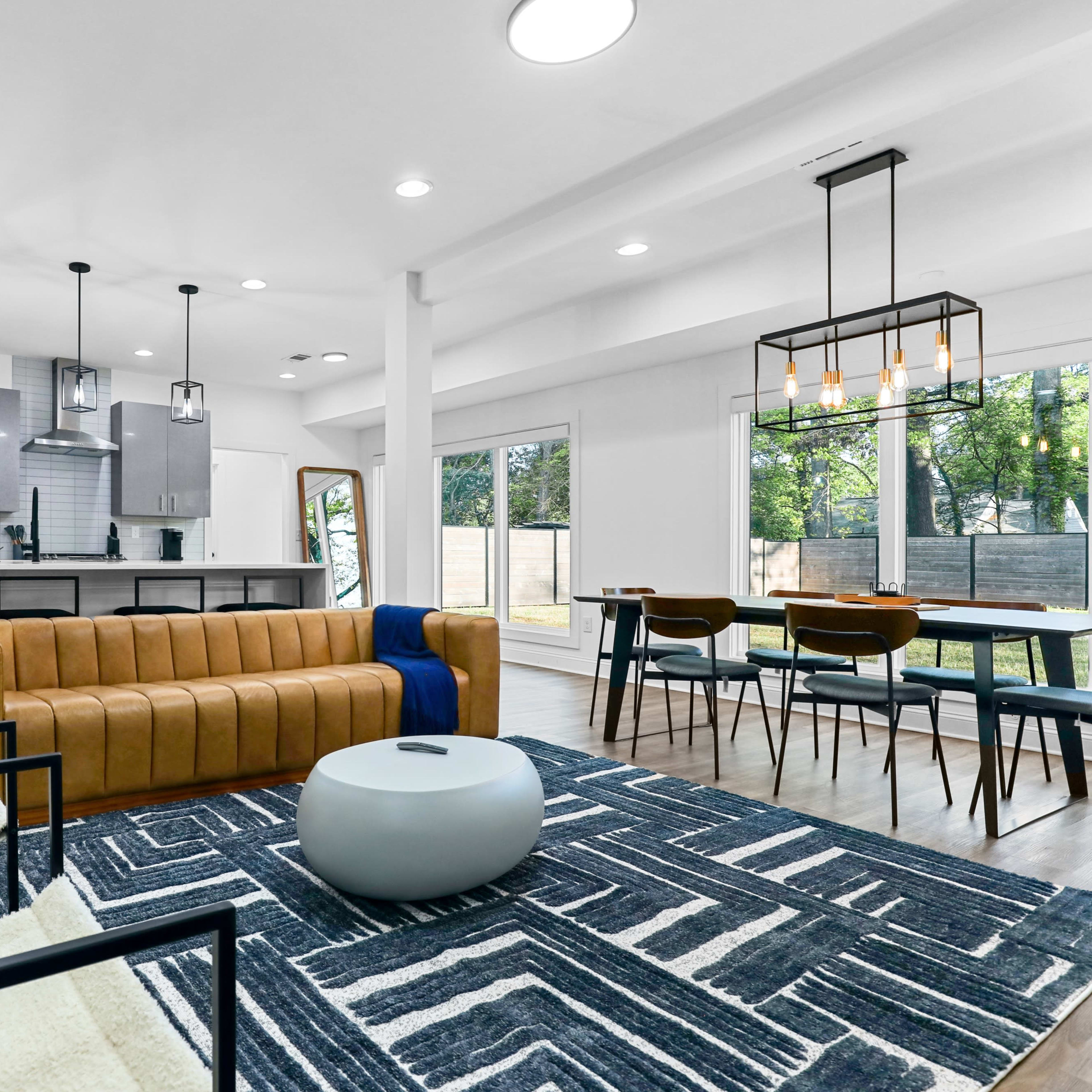 A modern living space with a brown leather couch, black chairs, a patterned rug, and a dining area with a wooden table and chairs, all set in front of large windows overlooking a yard.