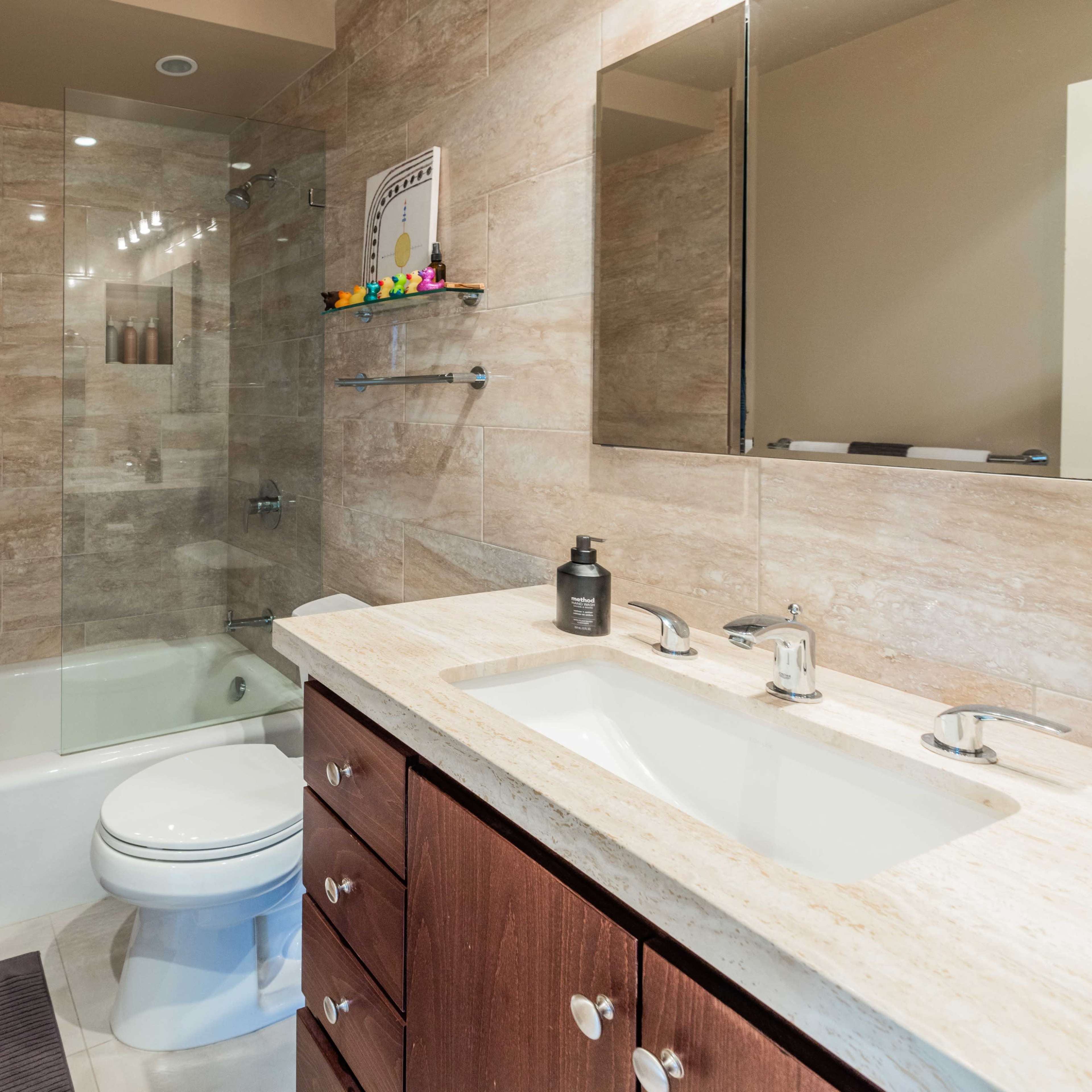 A modern bathroom featuring a glass-enclosed shower, a bathtub, a double sink vanity with wooden cabinetry, and tiled walls.