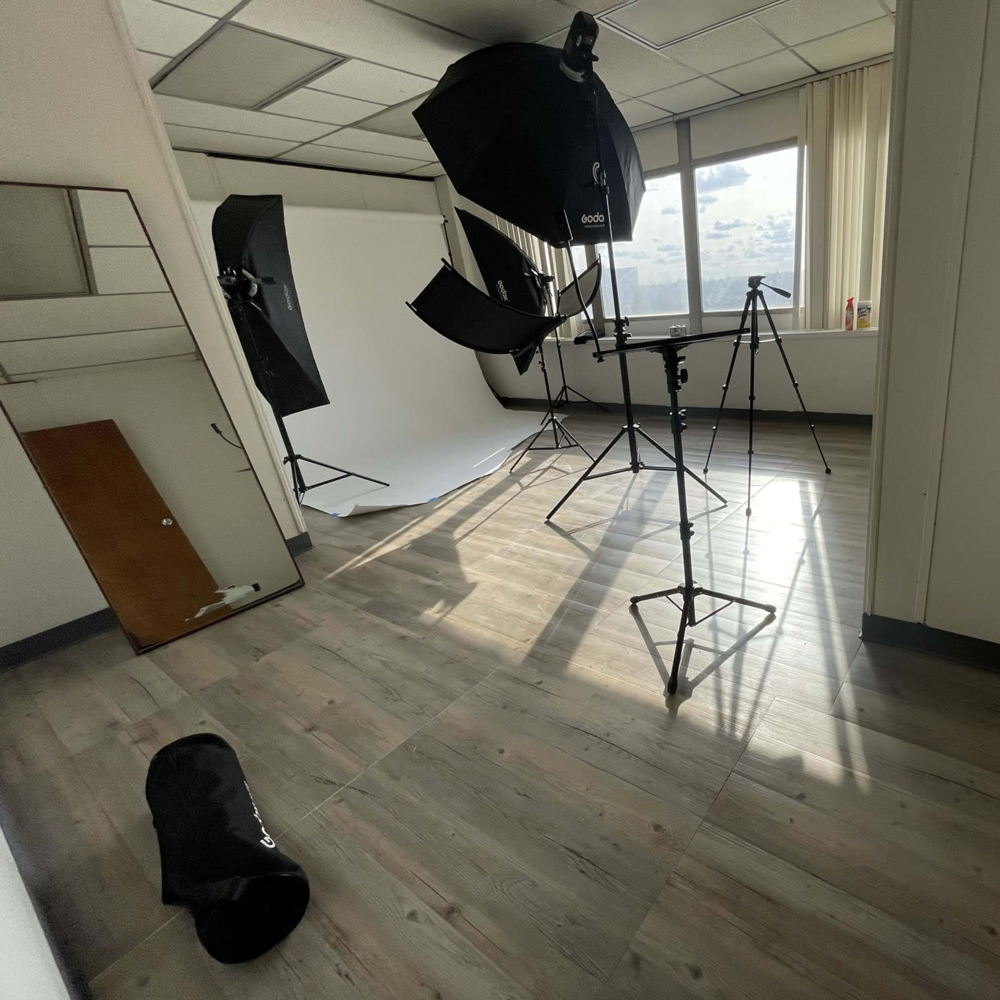 A photography studio with various equipment including softboxes, a tripod, and a backdrop, set up on a hardwood floor.