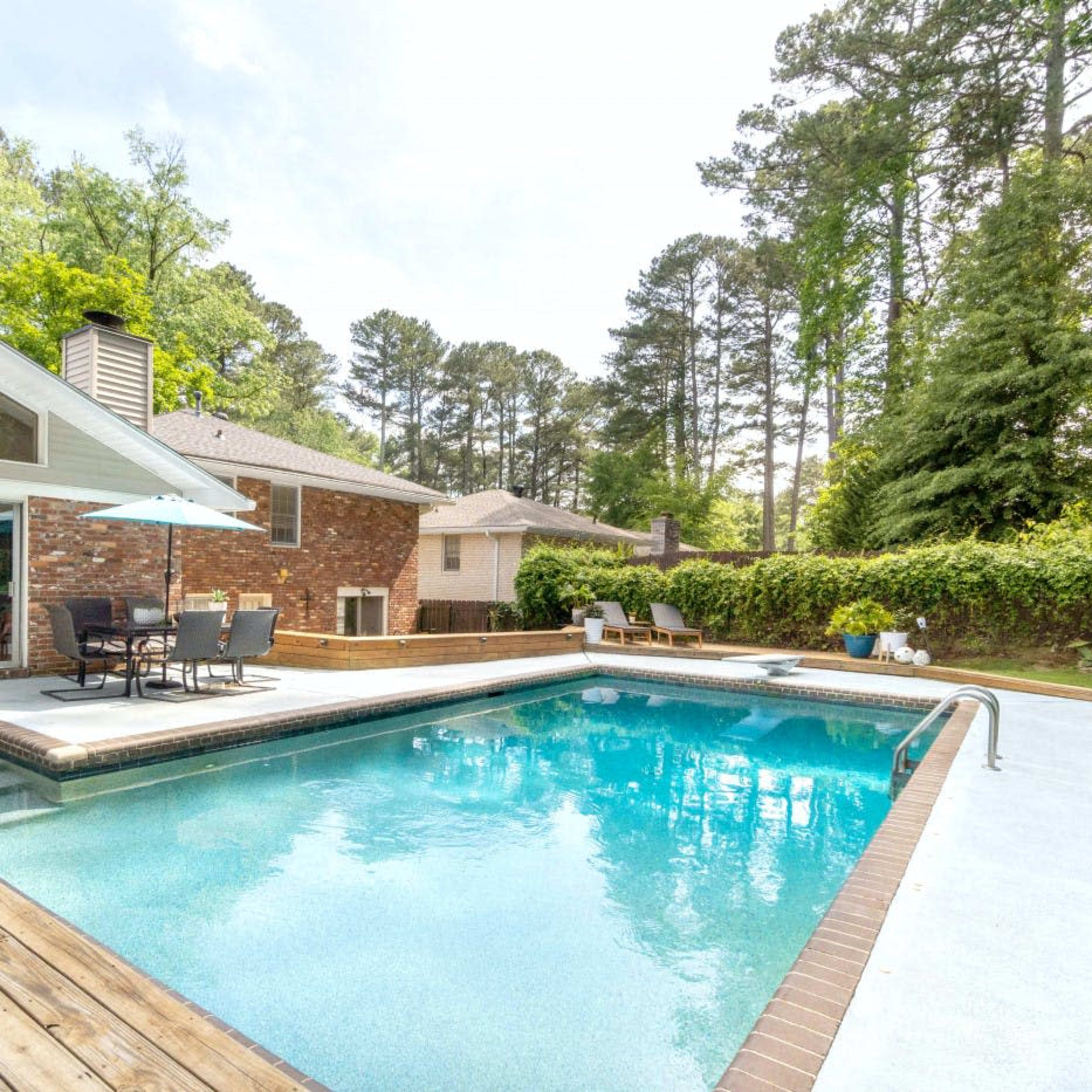 A backyard swimming pool surrounded by wooden decking, with a patio area and tall trees in the background.