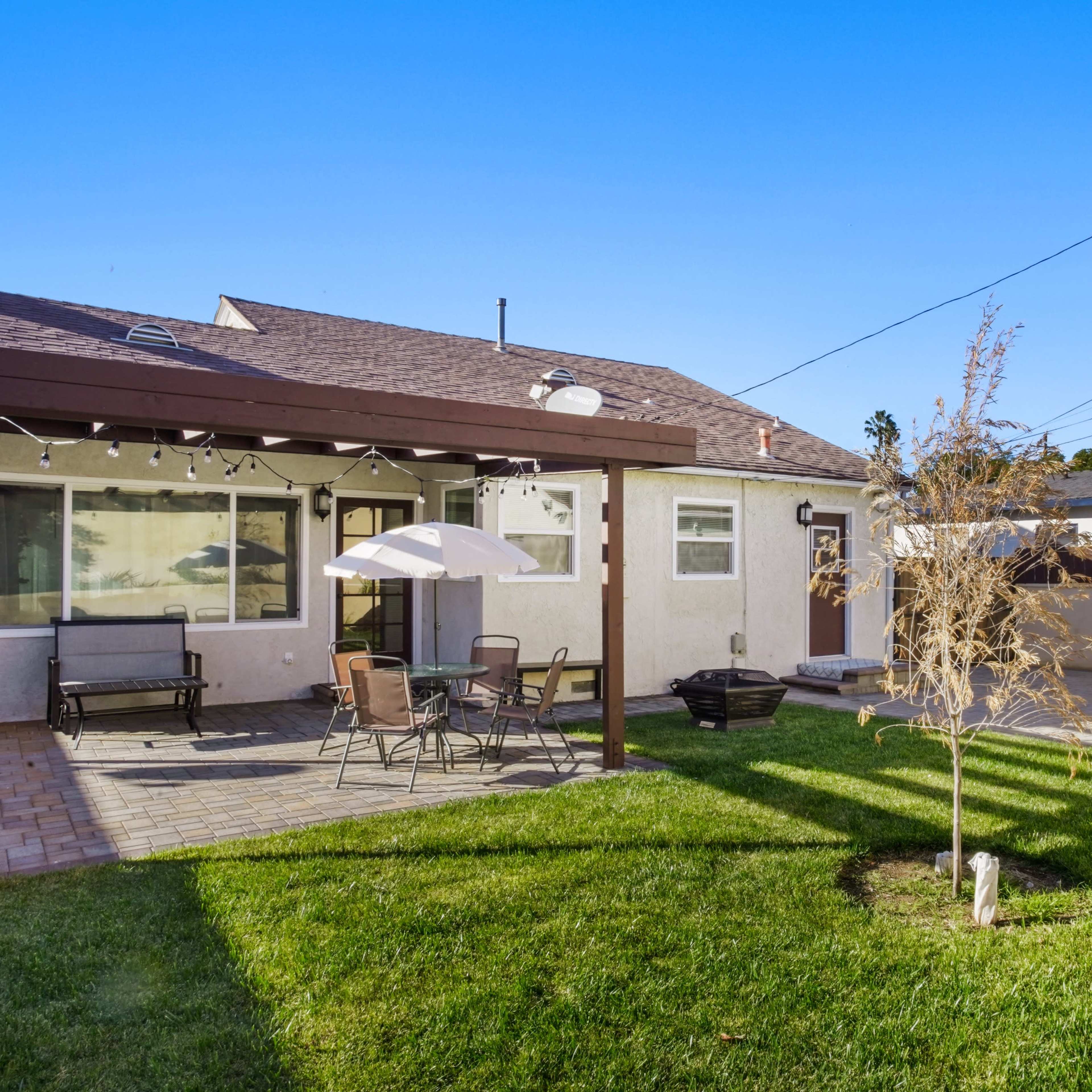 A backyard with a patio area featuring a table and chairs, an umbrella, and a small tree surrounded by grass, along with a storage shed visible in the background.