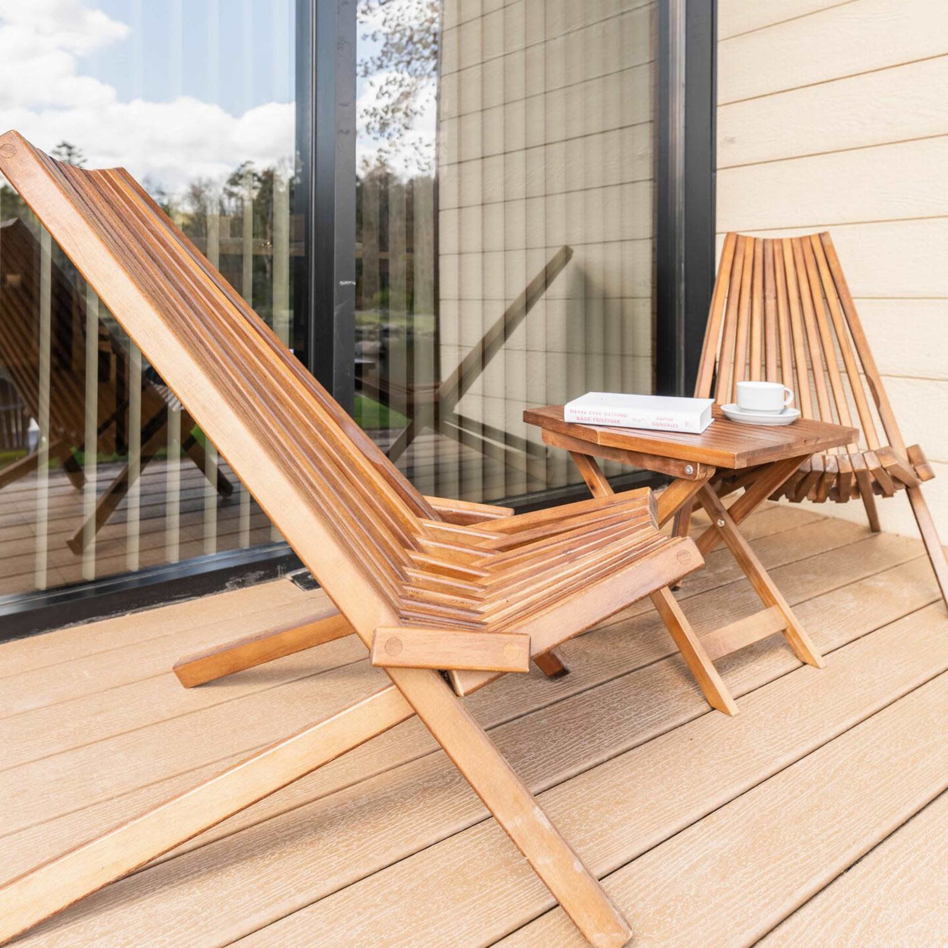 A pair of wooden lounge chairs and a small table are positioned on a deck, adjacent to large glass windows that overlook the outdoors.