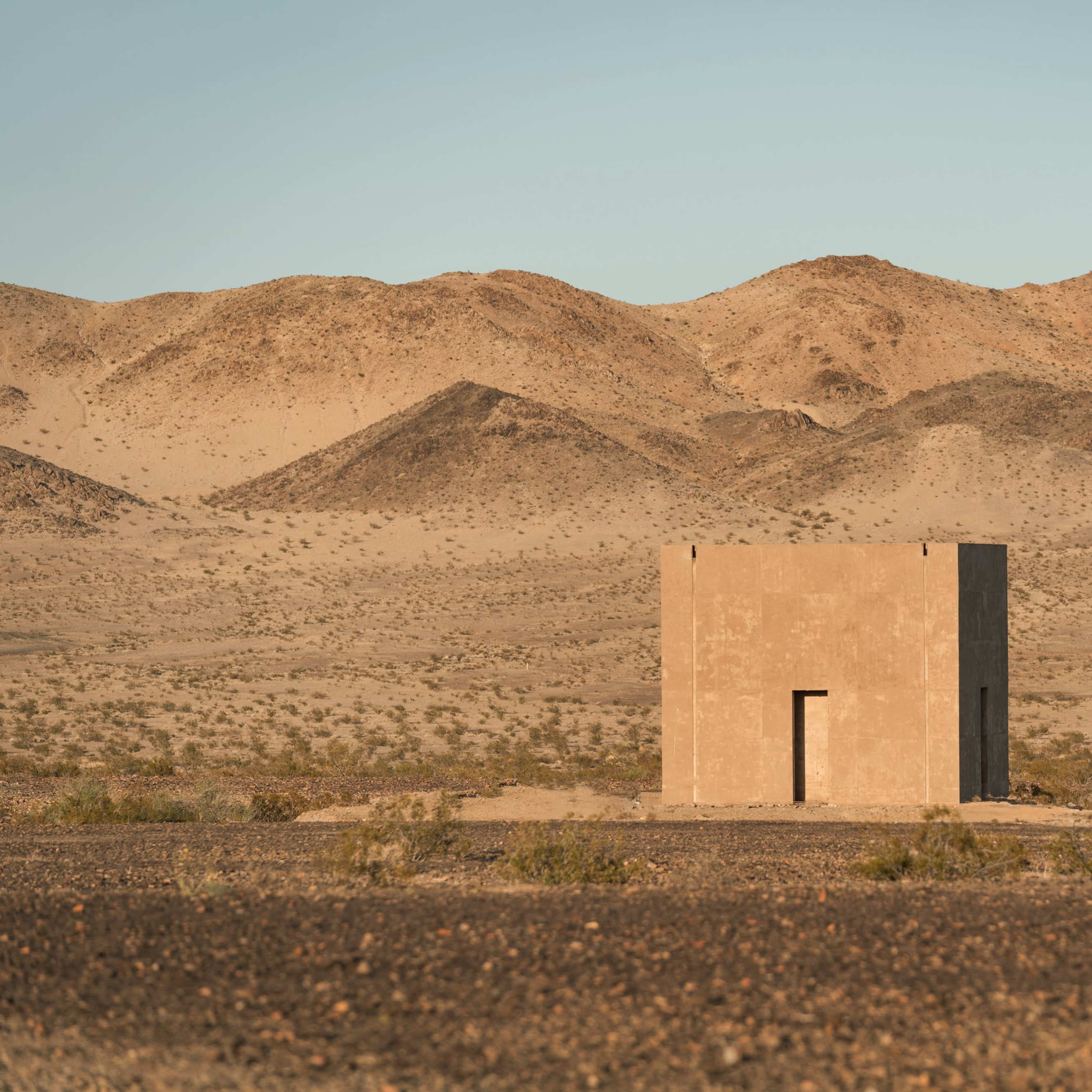 A simple, square building stands alone on a barren landscape surrounded by mountains and desert.