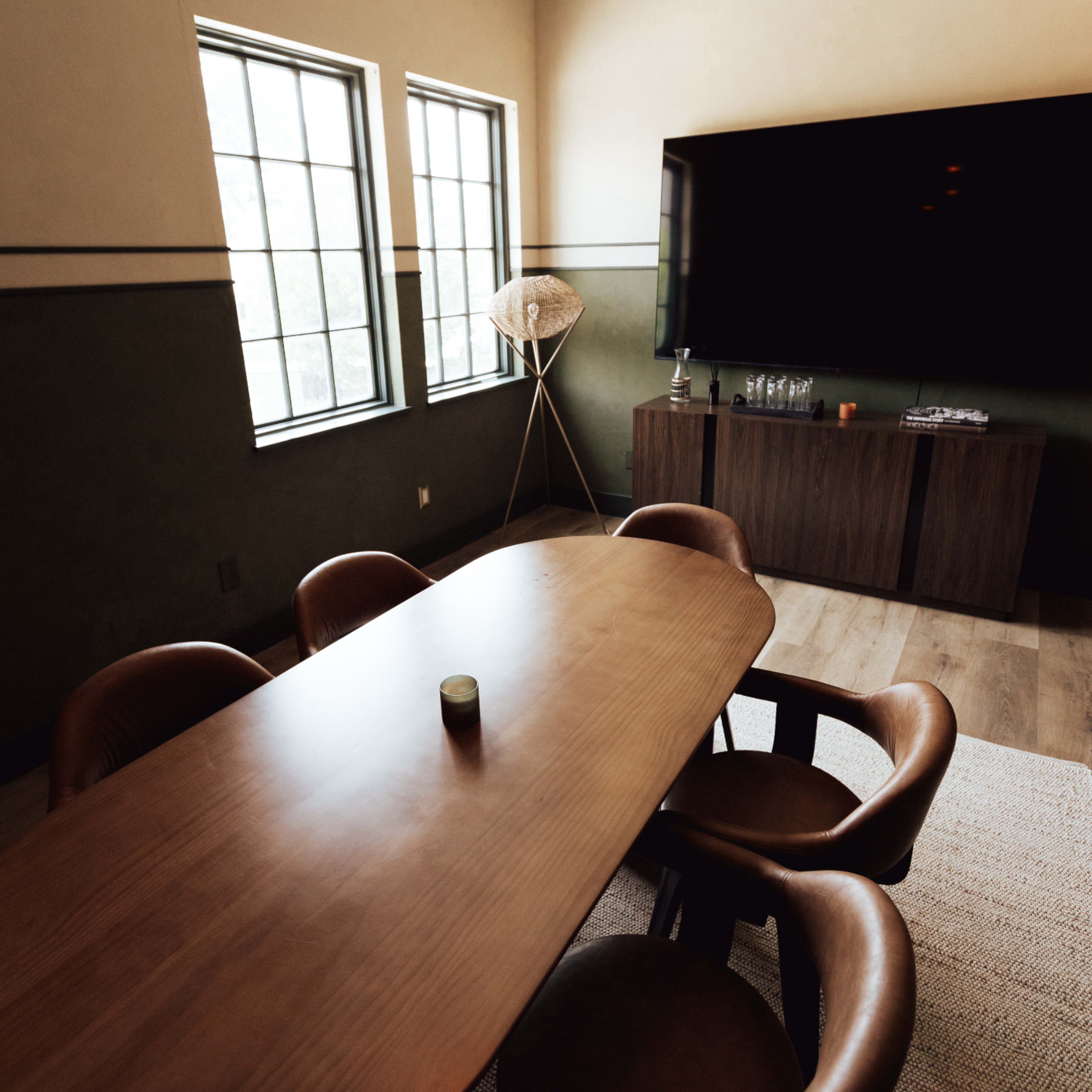A rectangular wooden dining table with circular chairs is set in a room featuring two windows, a television, and a floor lamp.