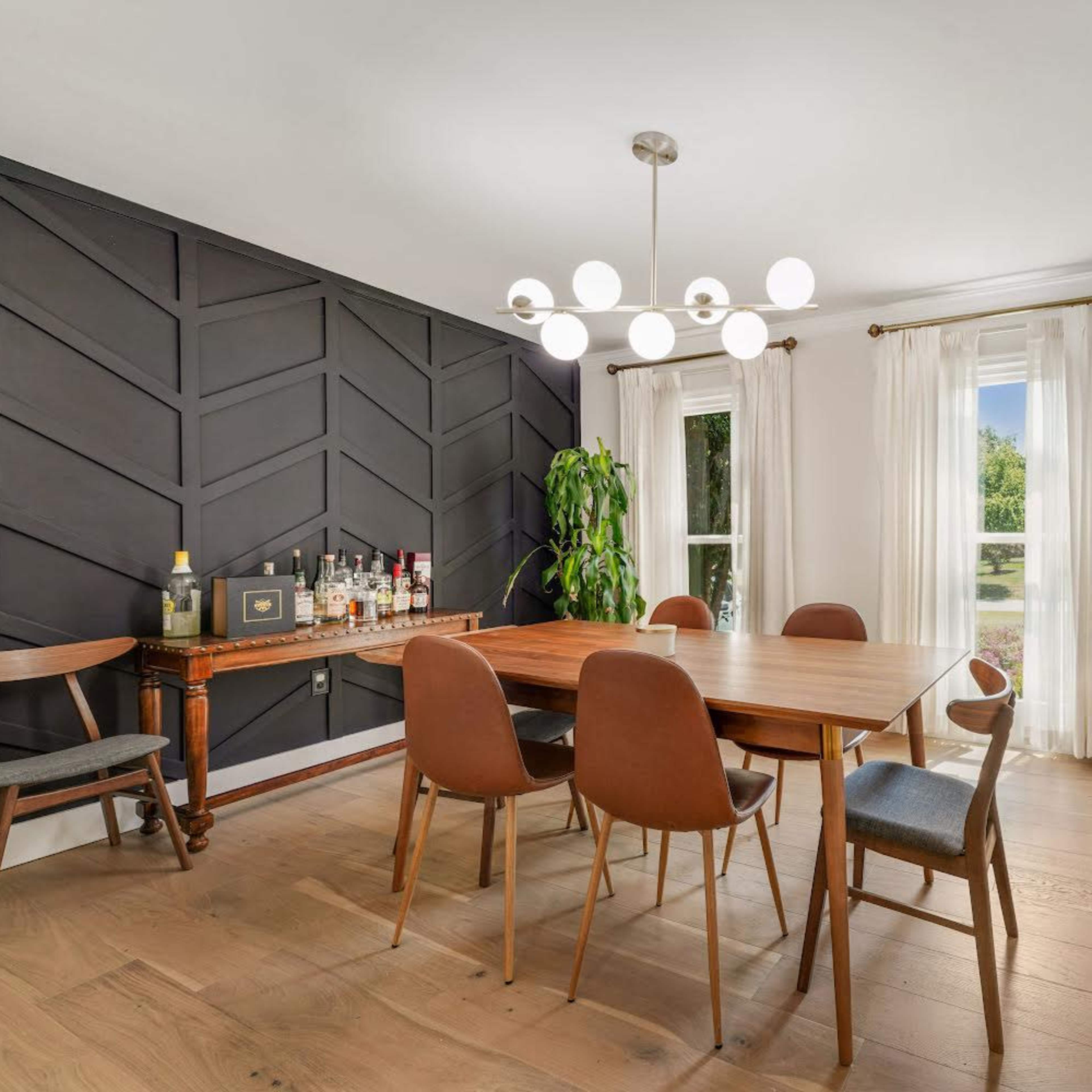 A dining room with a wooden table surrounded by brown chairs, a black geometric accent wall, and large windows with sheer curtains.