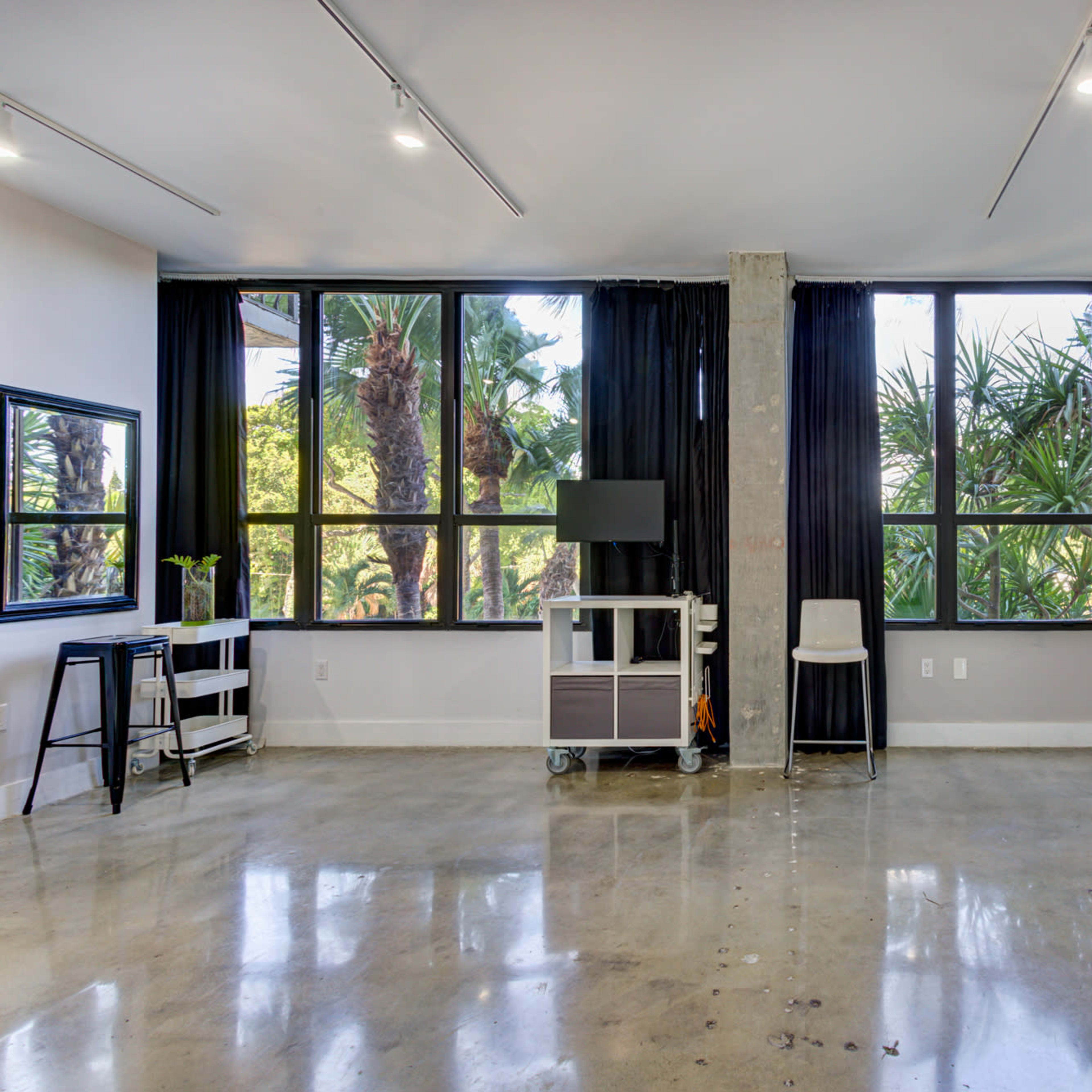 A spacious room with large windows revealing palm trees outside, featuring polished concrete floors and minimal furnishings.