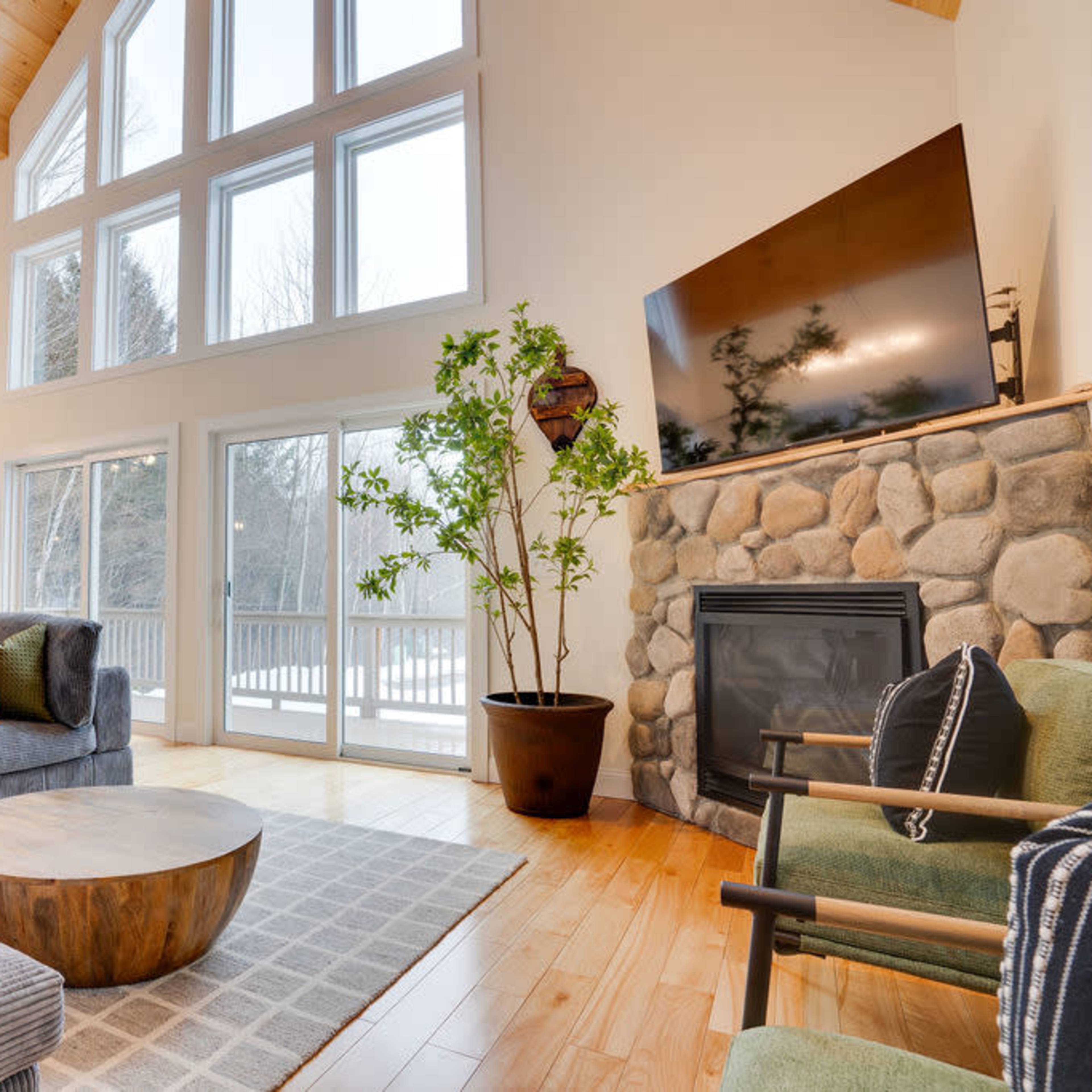 A modern living room with large windows, a stone fireplace, and a wooden ceiling, furnished with a gray sofa, a round wooden coffee table, and a green chair.