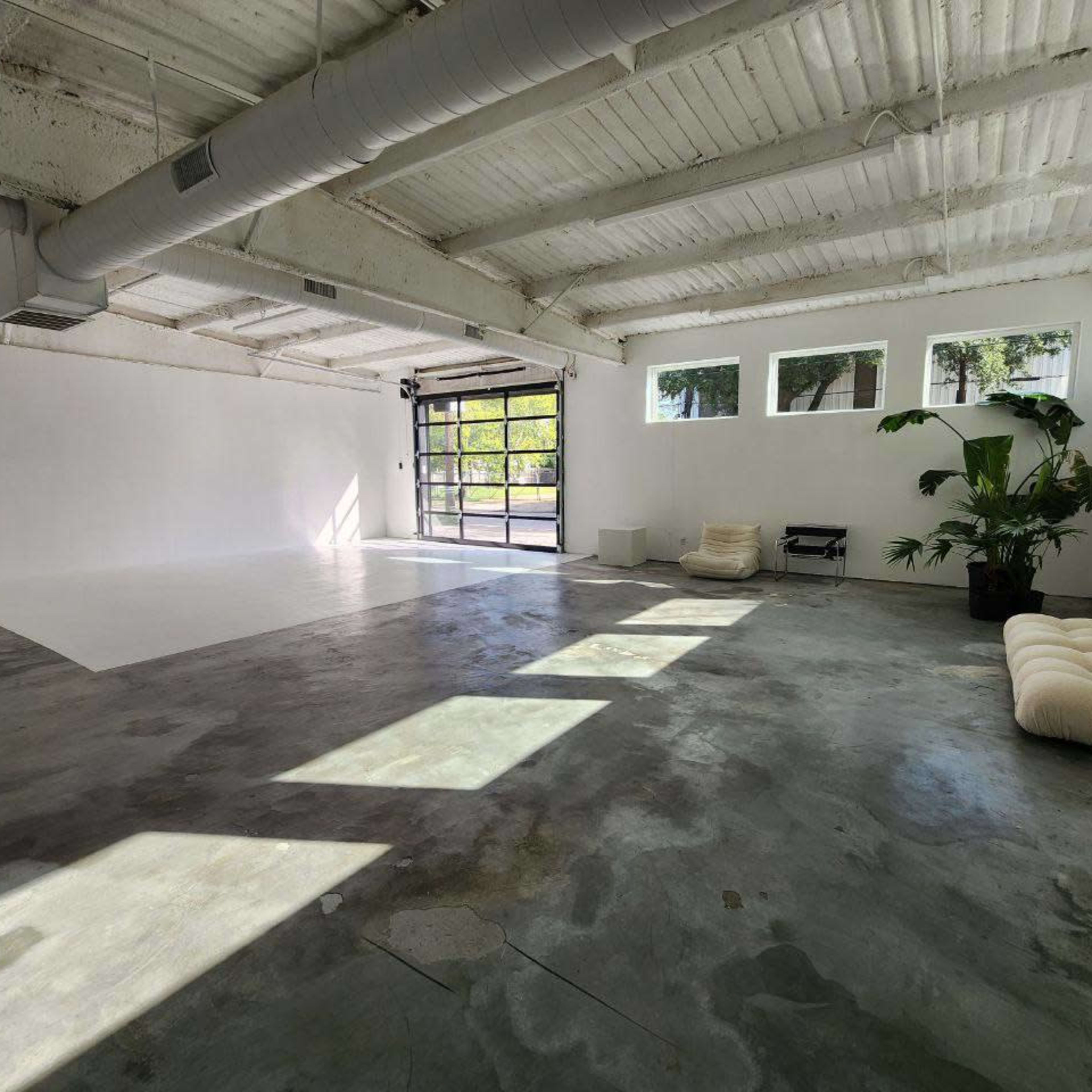 A spacious, empty room with concrete floors, large windows, and a garage-style door that opens to an outdoor area.