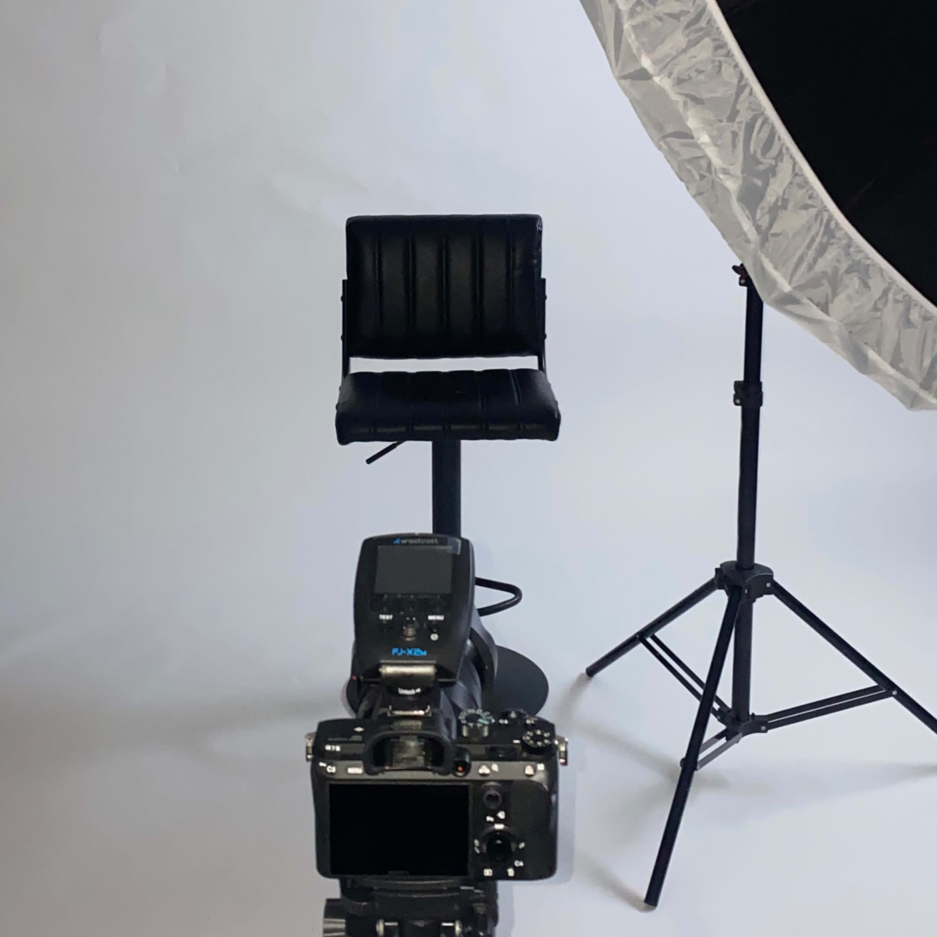 A black stool is positioned in front of a camera and a large softbox light against a plain backdrop.