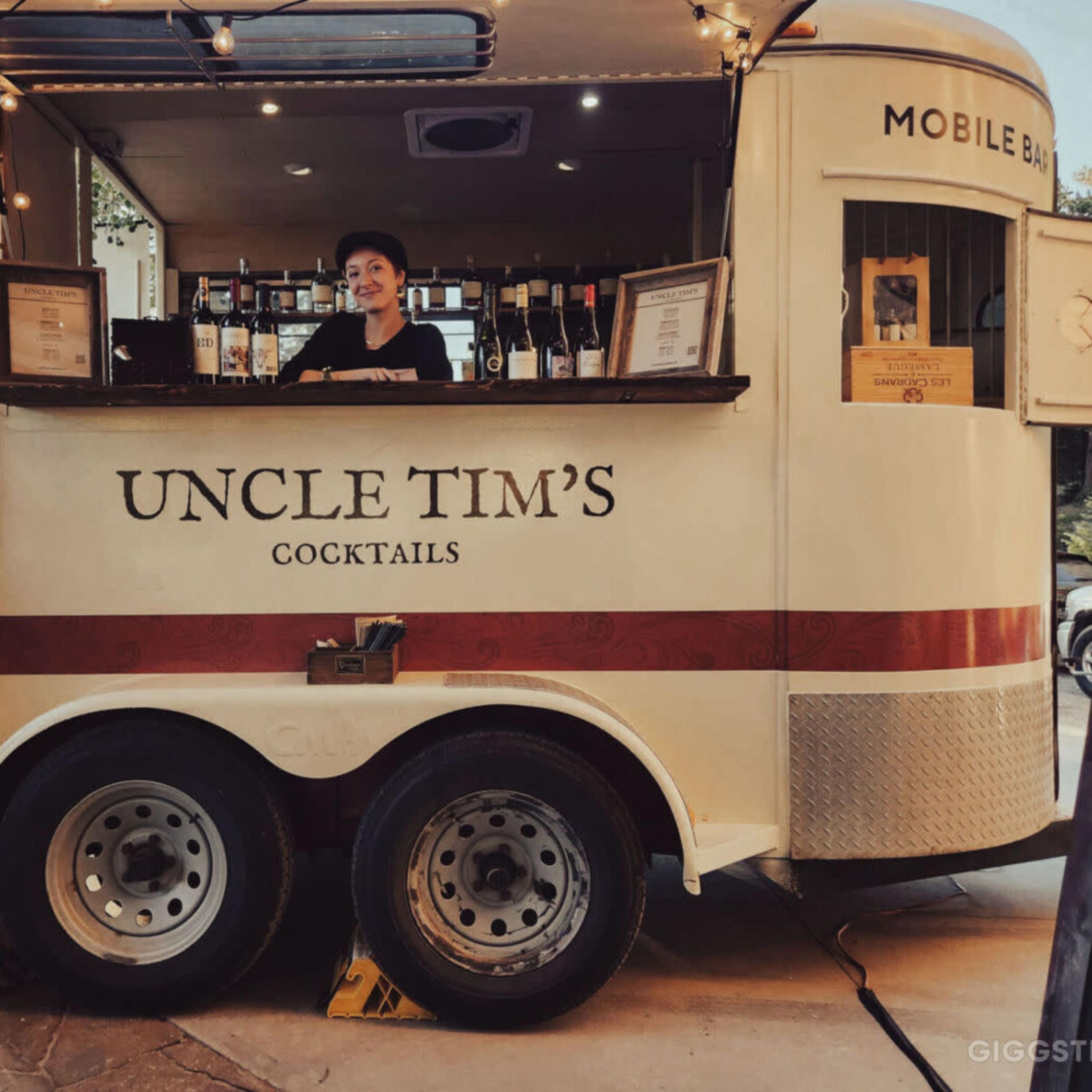 A mobile cocktail bar named "Uncle Tim's" is set up, featuring a bartender and a selection of drinks behind a wooden counter.