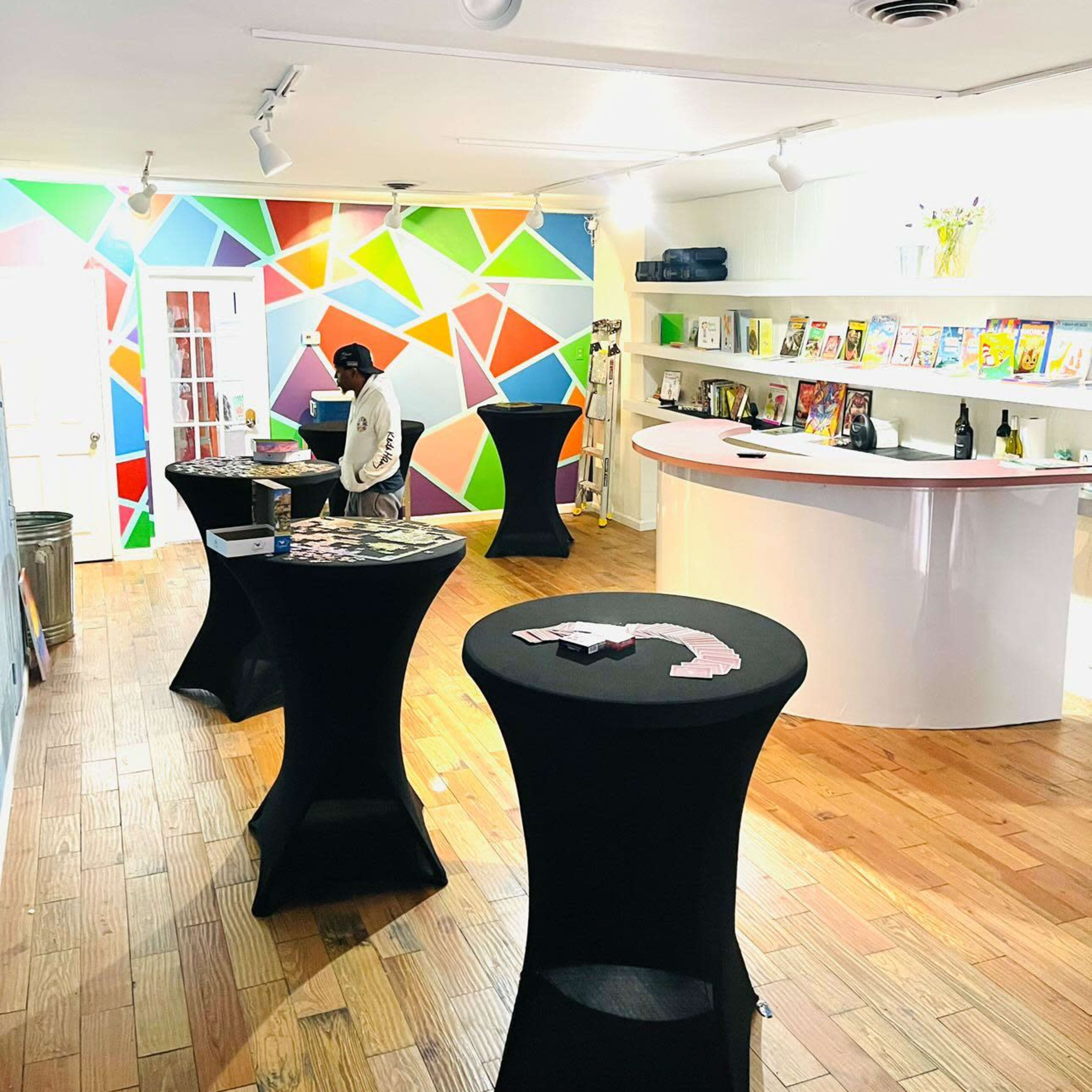 A brightly colored room with geometric wall patterns, four high tables with black covers, and a person sitting at one of the tables.