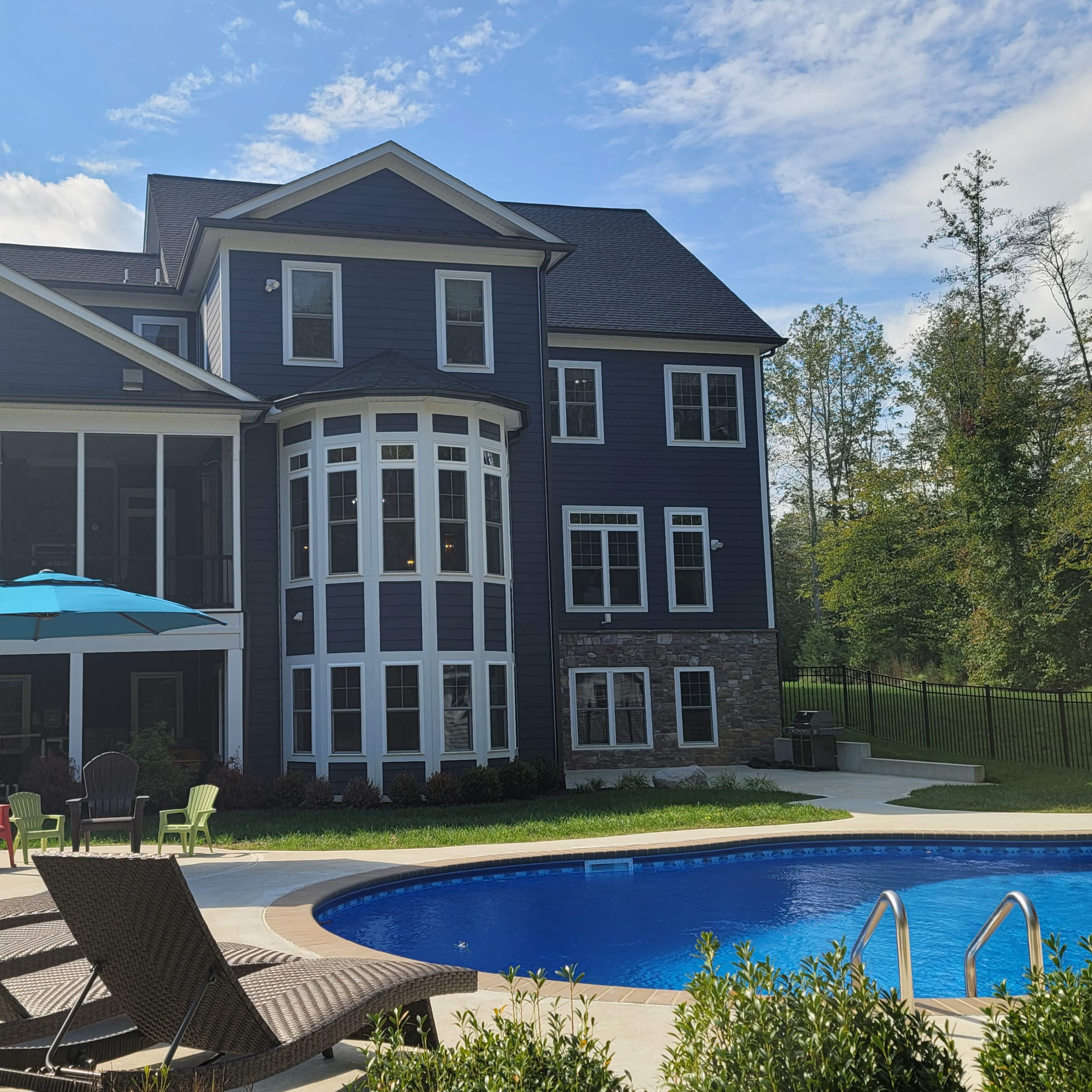 A large, modern house with a stone and dark siding exterior, a swimming pool in the foreground, and a patio with lounge chairs and an umbrella.