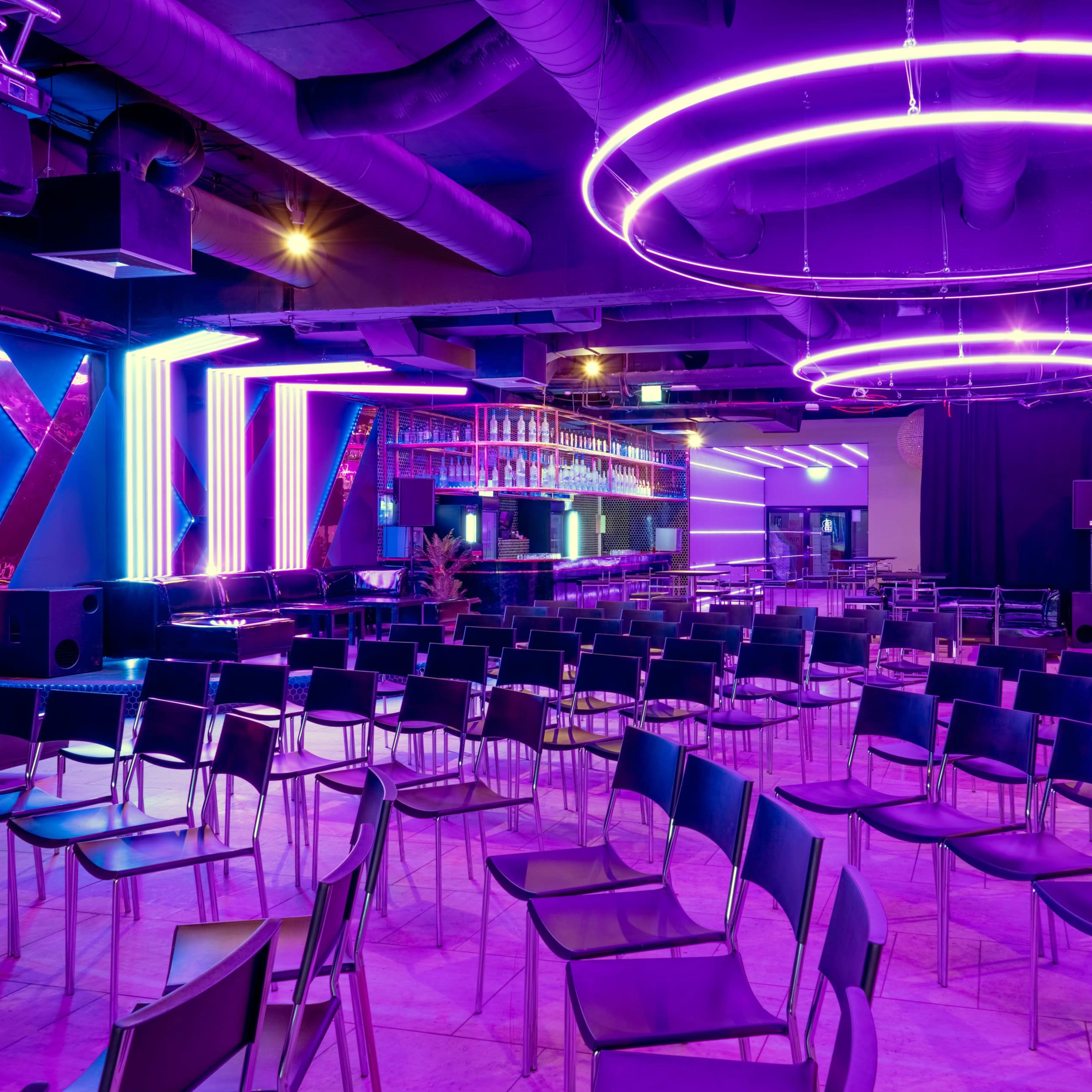 A modern setting with rows of black chairs arranged in front of a bar area, featuring colorful neon lights and a stage backdrop.