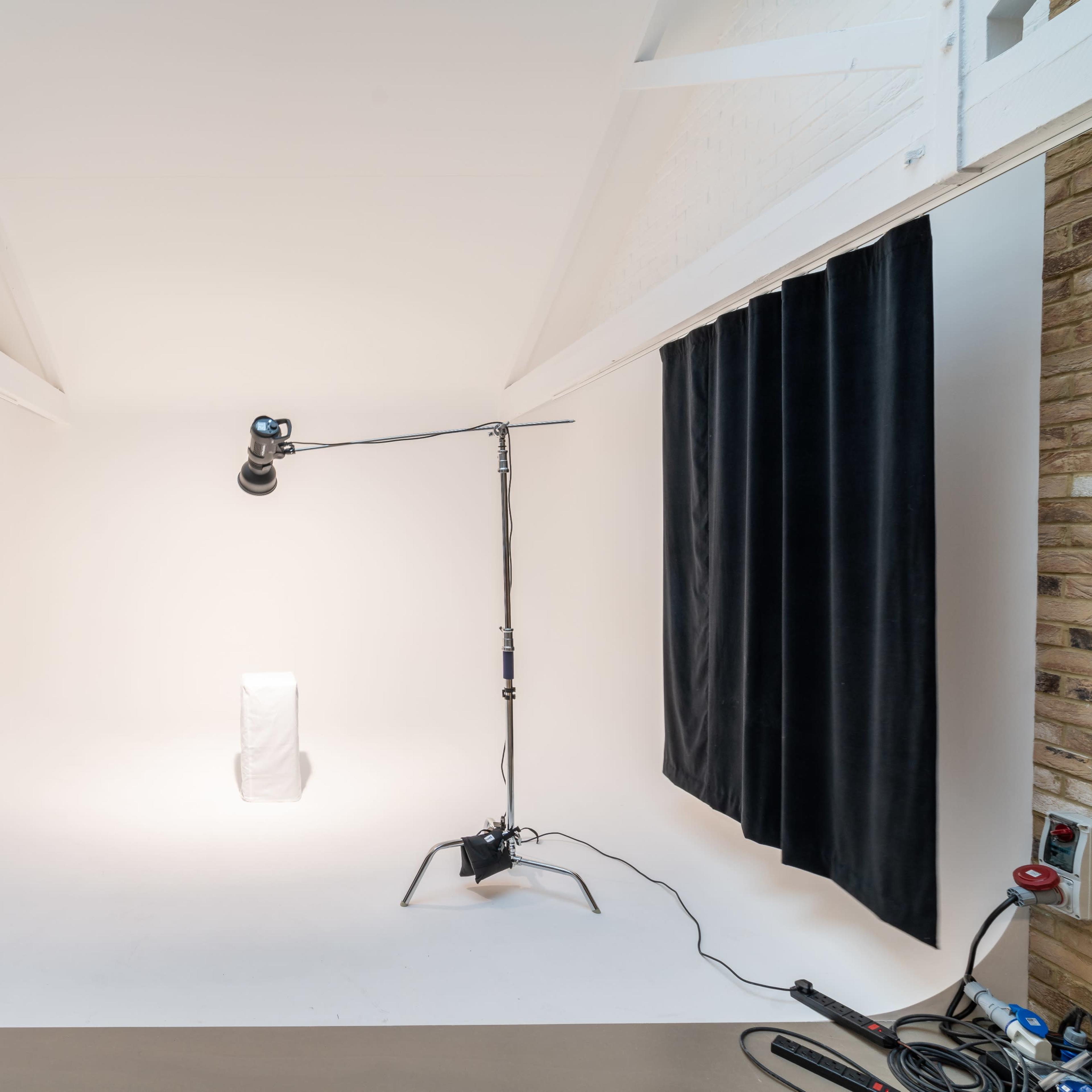 A photography studio with white walls, dark curtains on two sides, and a light stand positioned over a blank pedestal.