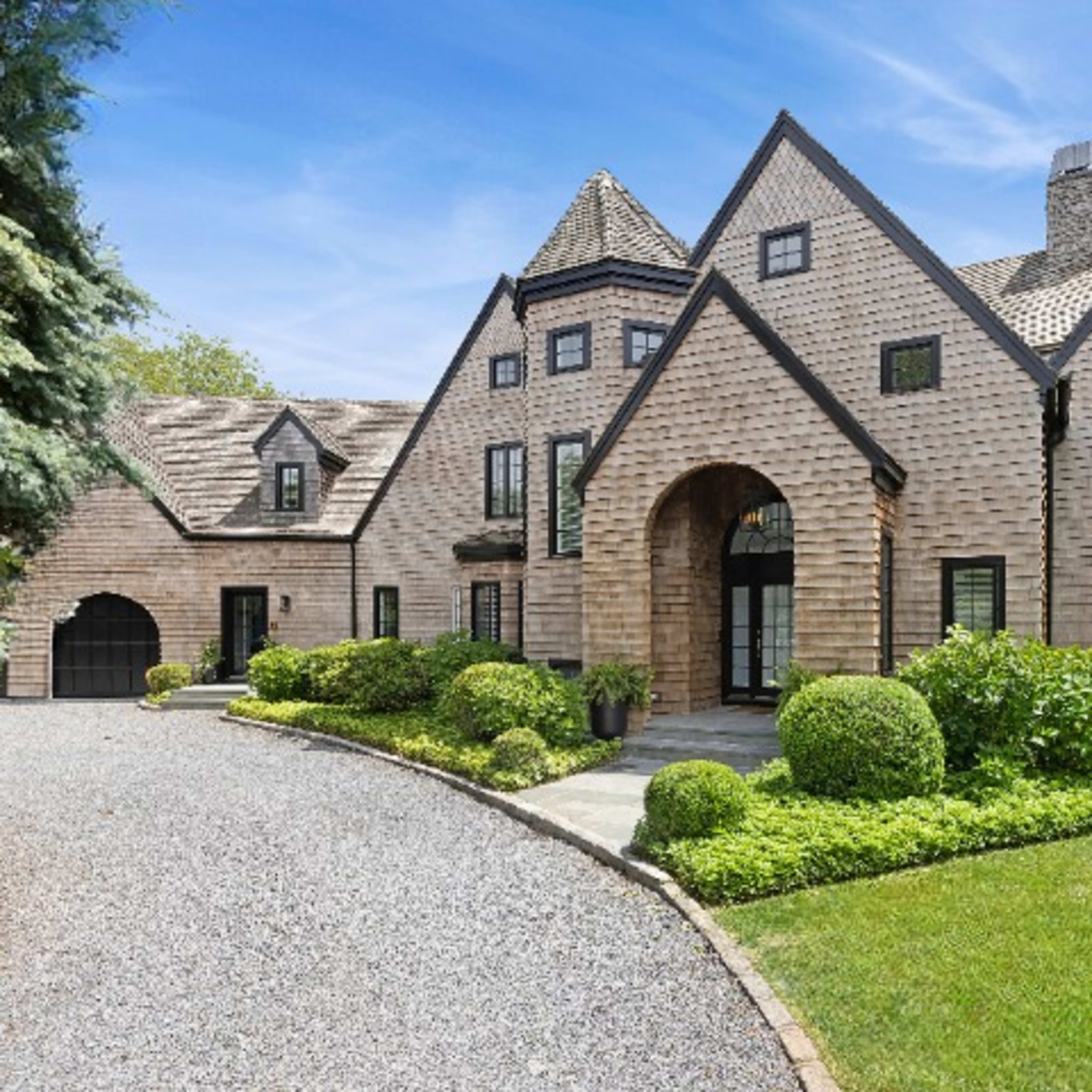 A large, multi-gabled house with a stone entrance and landscaped grounds is set along a gravel driveway.
