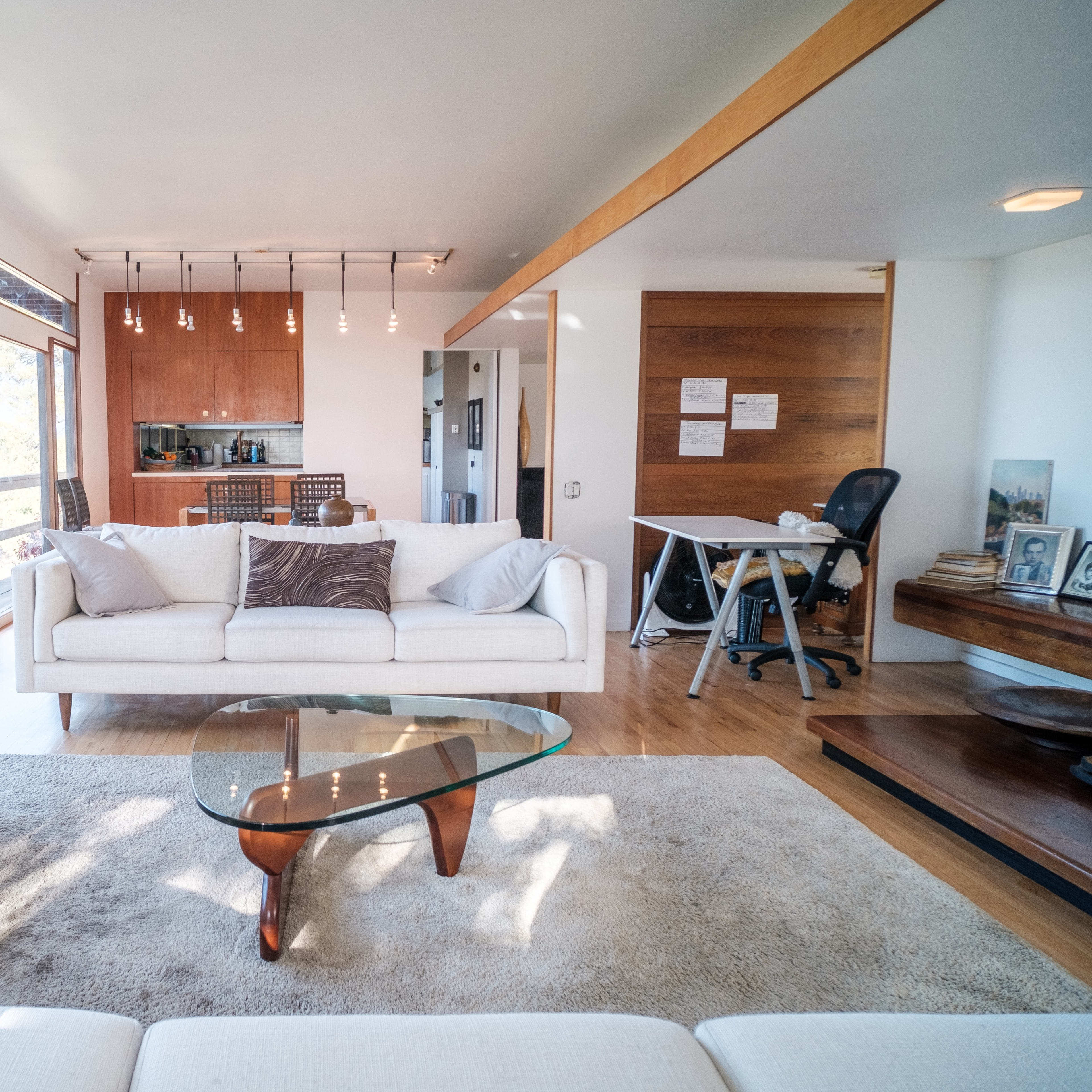A modern living room with a white sofa, a glass coffee table, and a view of a kitchen area in the background.