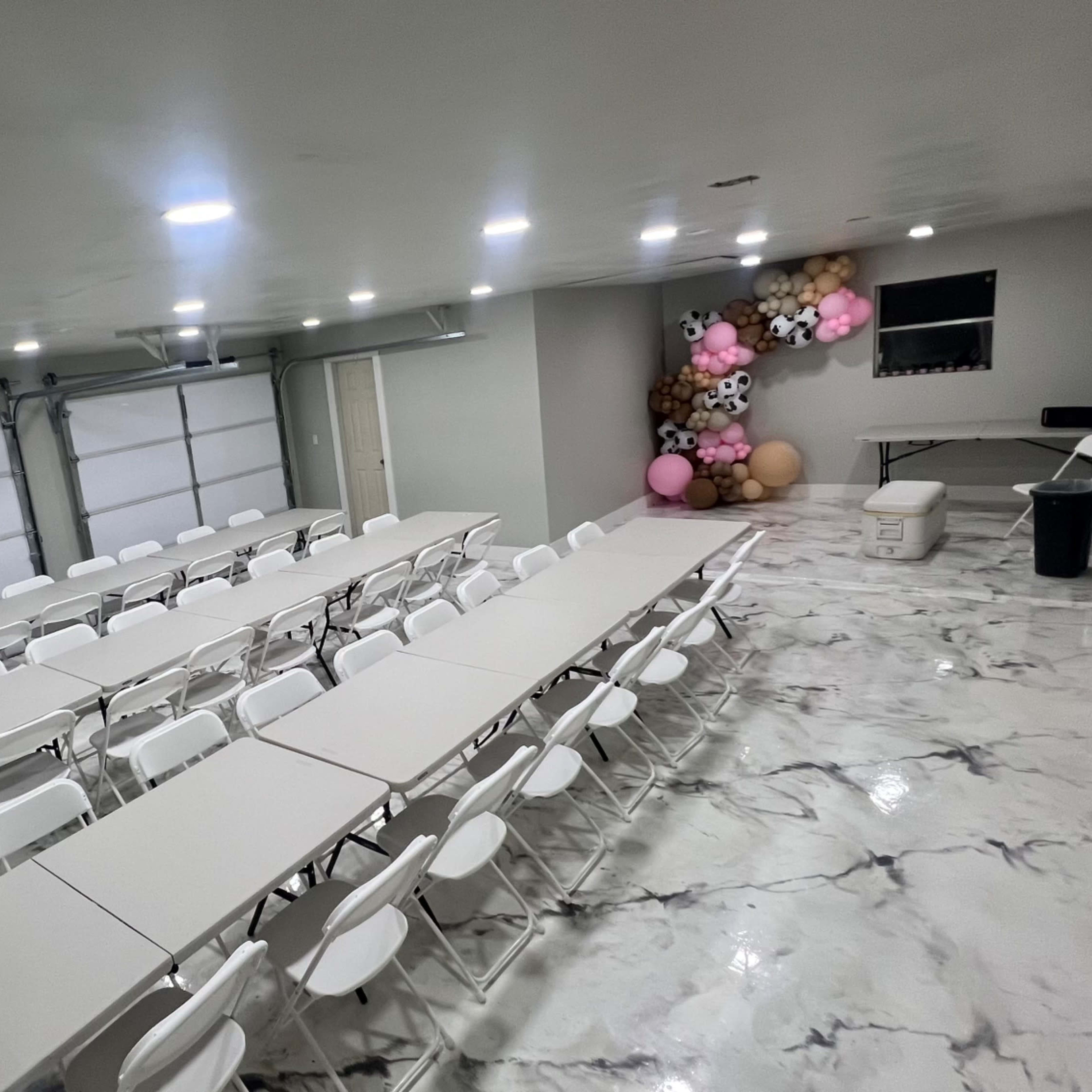 A spacious room with multiple long tables and chairs set in rows, featuring a wall adorned with decorative balloons.
