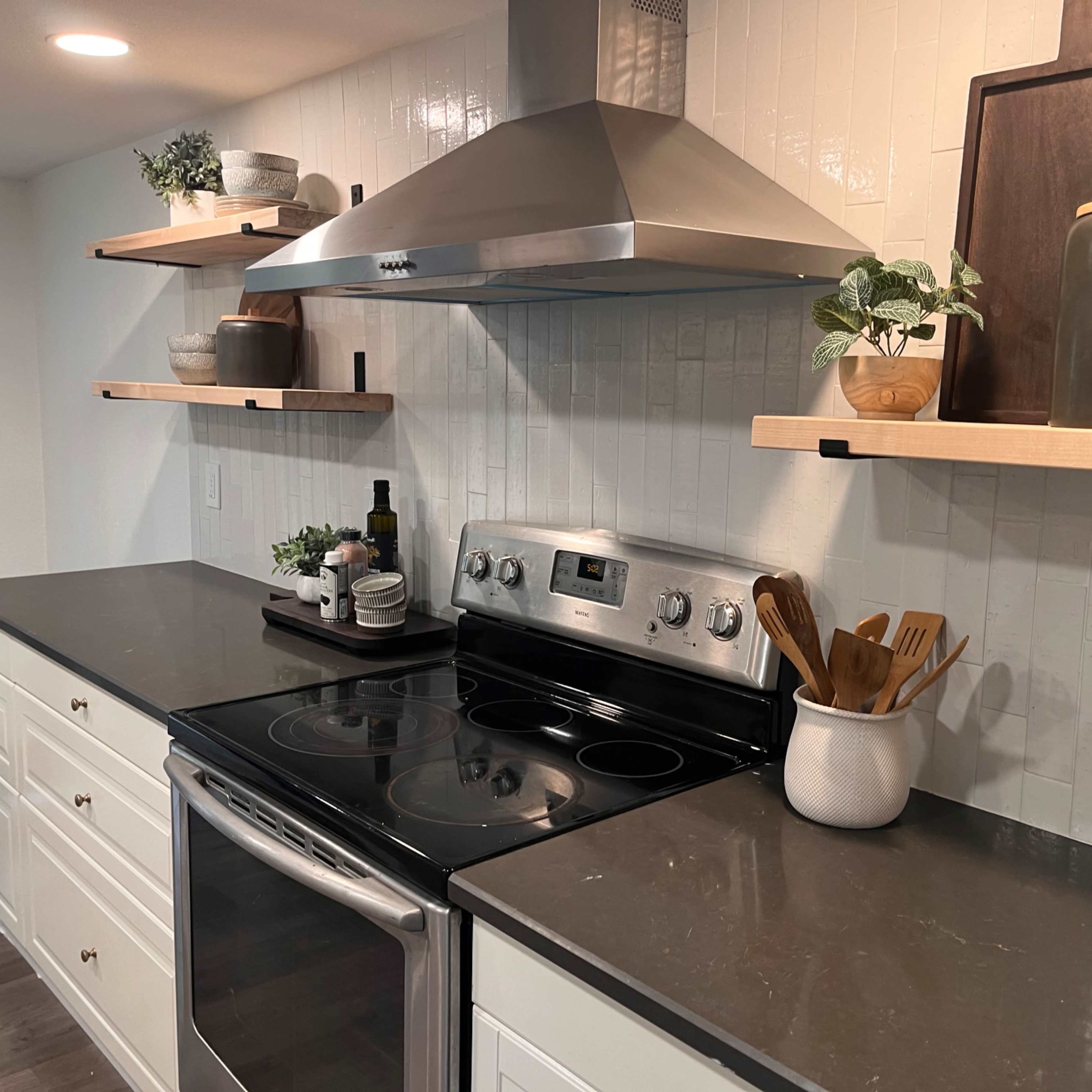 A modern kitchen with a stainless steel stove, a range hood, and wooden shelves displaying kitchenware and plants.
